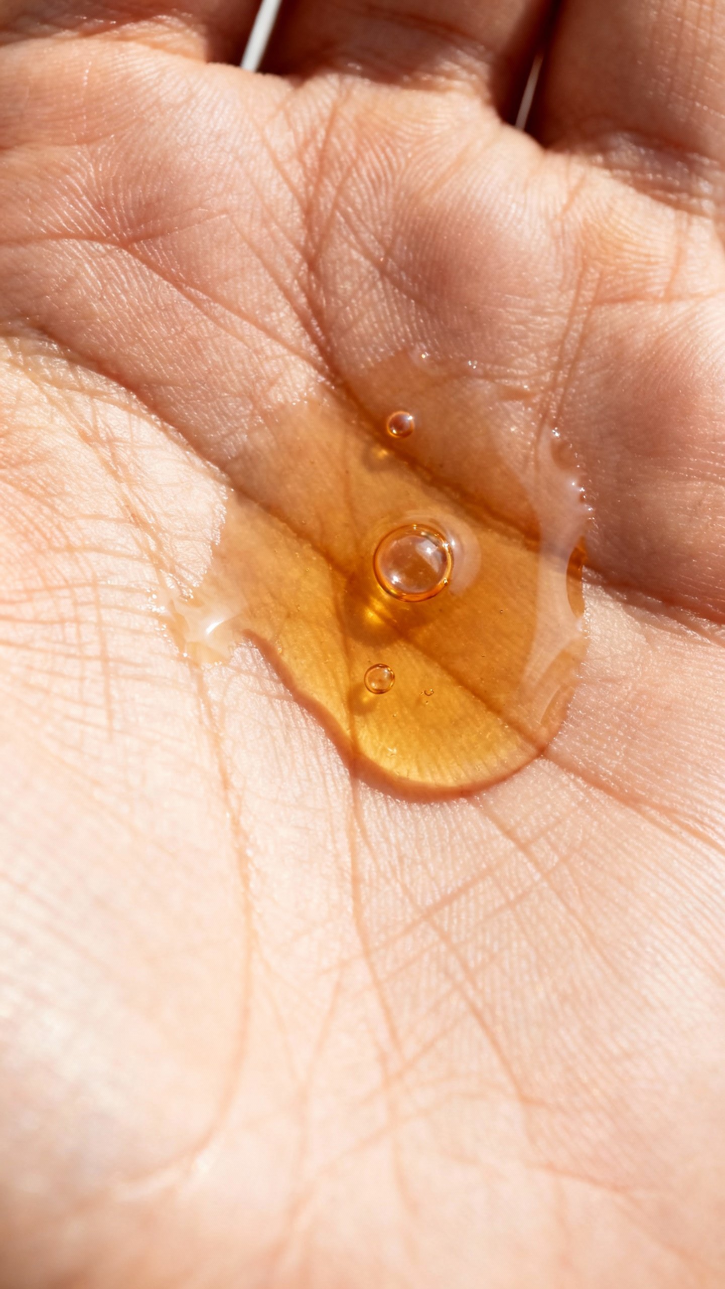 Overhead detail of face oil layering: clear hyaluronic droplets, amber squalane, on palm lines, soft