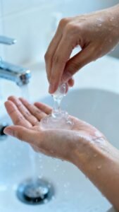 Hands applying clear gel cleanser to wet face, bathroom sink closeup
