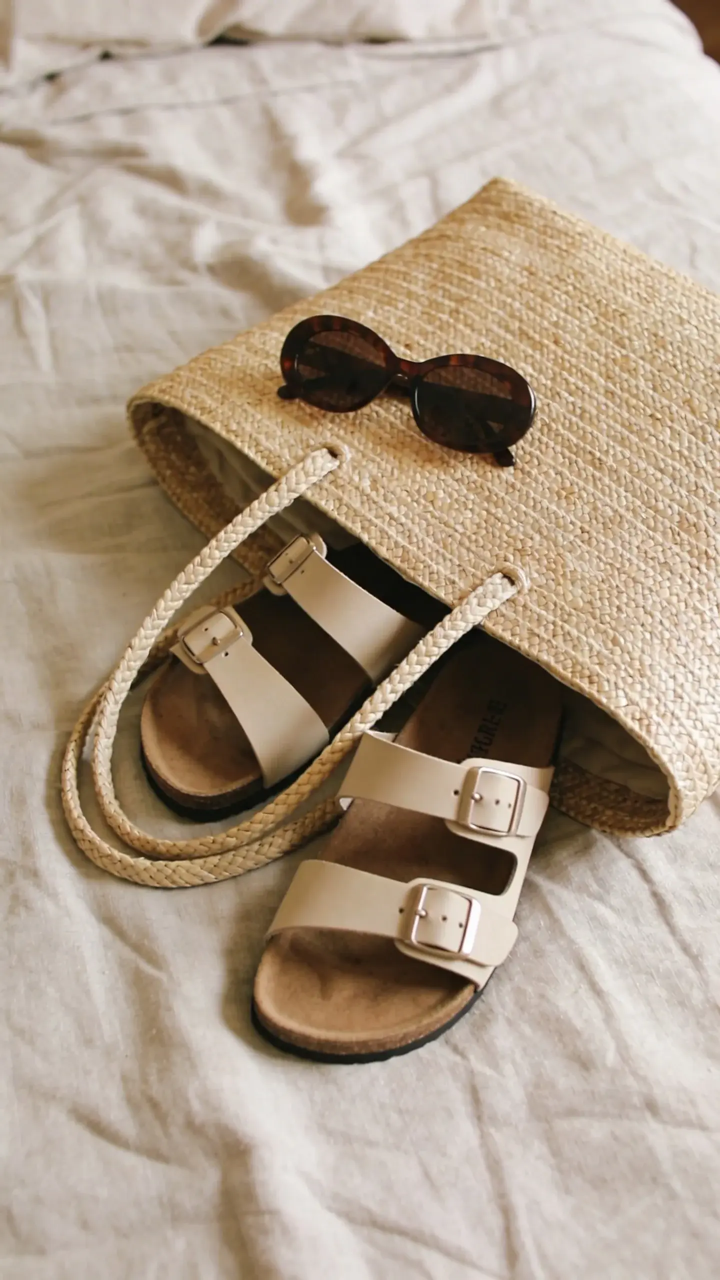 straw tote with sandals and sunglasses on linen bed