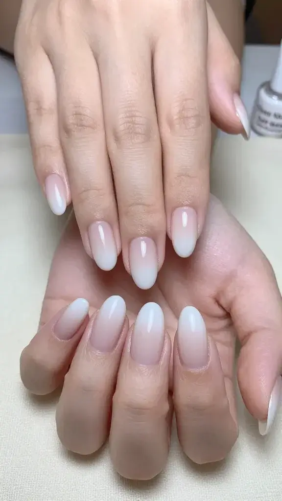 Top-down, ultra-realistic close-up photo of both hands resting lightly on a soft ivory linen surface, showcasing short-to-medium almond-shaped nails with a soft-blur “baby boomer” ombre: sheer neutral nude at the cuticle melting seamlessly into a whisper-white tip with an airbrushed, cloudlike gradient. The finish is glossy and glass-smooth with subtle natural reflection, cuticles clean and hydrated, natural skin tone with faint skin texture and soft micro-hairs visible for realism. Include a faintly visible makeup sponge edge in the background and a slim non-latex cleanup brush with a tiny hint of acetone sheen, plus two small polish bottles labeled “Sheer Nude” and “Soft White” slightly out of focus. Lighting is bright, diffused daylight from the left, gentle shadows, high-resolution macro detail; neutral color palette, no glitter, no decals, no harsh lines. Composition centered on nails, depth of field shallow for creamy bokeh, editorial beauty photography style.