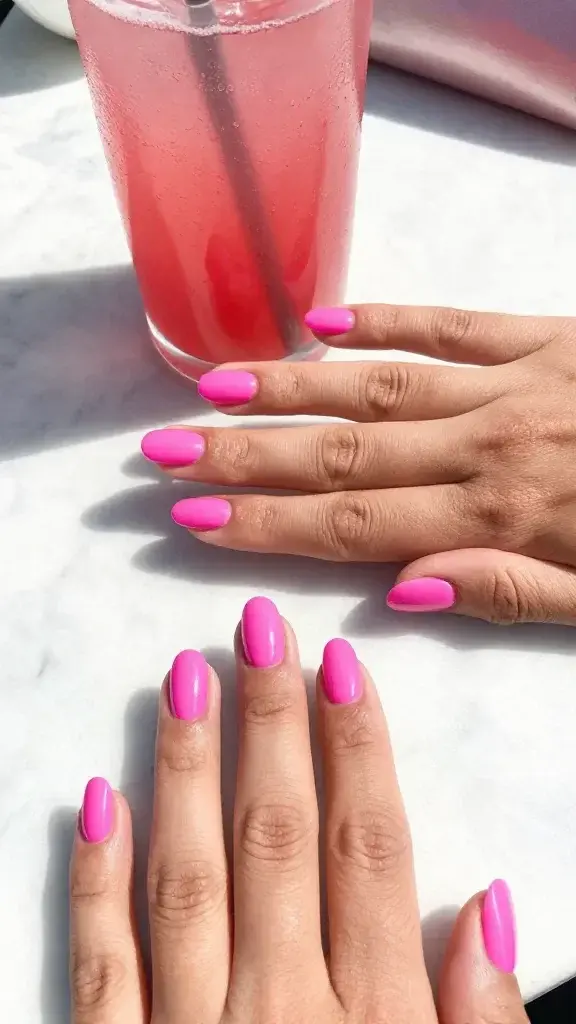 Top-down, ultra-realistic studio photo of both hands resting lightly on a sunlit white marble tabletop, showcasing a fresh Barbiecore punch pink manicure: short to medium oval nails painted in a crisp blue-leaning fuchsia (cool-toned hot pink) with a high-shine, vinyl-like finish. Two thin, even coats create saturated color with perfectly capped tips visible at the edges; no streaks, no bubbling, ultra-gloss reflection highlights. Natural, moisturized cuticles, subtle hand veins, soft skin texture, and a faint summery glow. Include a chilled fuchsia cocktail with condensation and a slim silver straw at the frame’s edge, plus a hint of a metallic pink clutch blurred in the background to suggest rooftop happy hour. Bright, diffused daylight from upper left casts soft shadows; color accuracy prioritized to emphasize the cool fuchsia tone that flatters all undertones. High-resolution macro detail on nail surfaces and tip capping; realistic depth of field keeps nails tack-sharp while background props remain softly out of focus.
