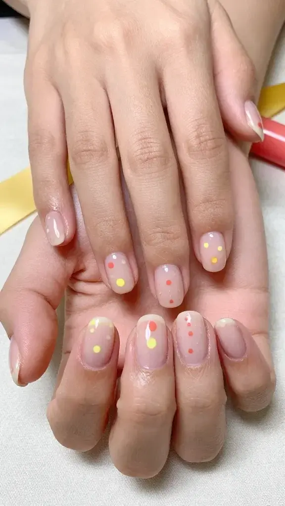 Top-down, ultra-realistic macro photo of both hands resting on a soft linen surface, showcasing short, neatly filed natural nails with a glossy sheer nude-pink base. Each nail features minimalist sunshine micro-dots in warm tones: a single tiny canary-yellow dot perfectly centered 2–3 mm above the cuticle on the thumbs, index, and pinky; two staggered micro-dots (one lemon yellow, one soft coral) on the middle fingers placed asymmetrically near the lower third of the nail; and a delicate diagonal line of three coral micro-dots on each ring finger starting from the lower inner corner moving upward toward the outer edge. Cuticles are clean and hydrated, skin tone is light-medium with warm undertones, subtle natural hand texture visible. Lighting is bright, diffused daylight for soft shadows and true color. Background includes a hint of a yellow silk ribbon and a coral lip balm tube out of focus to echo the palette. Camera: 50mm equivalent, f/4, high-resolution, color-accurate, editorial beauty photography style.