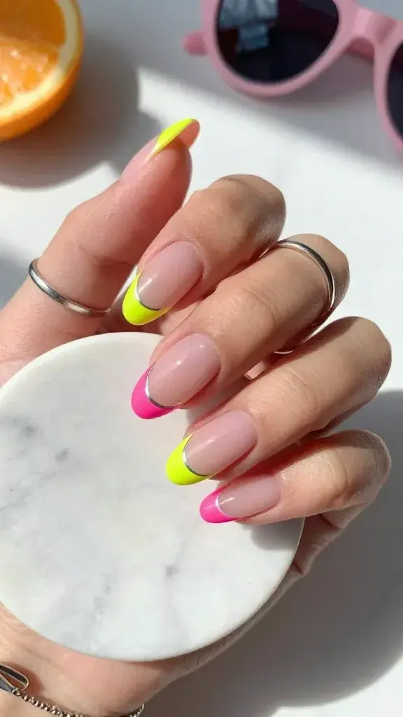 Top-down, hyper-realistic close-up photo of a single hand resting on a sunlit white marble surface, showcasing fresh “French tips, but electric” summer nails: short-to-medium almond shape with a sheer cool pink jelly base for natural contrast. Tips alternate in neon shades—highlighter yellow on index, hot pink on middle, neon lime on ring, and hot pink on pinky—with ultra-precise, ultra-thin curved French lines. Under each neon tip, add a razor-thin chrome silver micro-line that catches the light for a subtle metallic pop. Nails are glossy with high-shine top coat; cuticles are clean and hydrated. The hand wears a minimal silver stacking ring and a fine chain bracelet. Soft morning sunlight creates gentle shadows and a slight prismatic reflection on the marble, enhancing the neon brightness. Depth of field is shallow, focusing crisply on the nail edges and chrome lines; background softly blurred with a hint of a citrus slice and a pink sunglasses arm peeking into frame for a summery vibe. Photographic style: professional beauty editorial, natural color grading, no heavy filters, ultra-detailed texture of nail surface and skin.