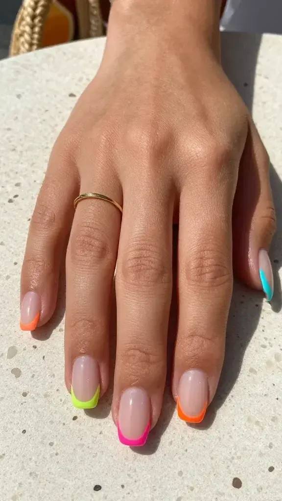 Top-down, ultra-realistic close-up photo of a single hand resting on a sunlit terrazzo tabletop, showcasing a micro French manicure with neon edges. Nails are medium-short, soft square shape with a sheer milky nude base (slight semi-transparency, subtle pink-beige warmth) and ultra-thin neon micro tips: index—neon lime, middle—electric coral, ring—hot pink, pinky—neon tangerine, thumb—neon turquoise. The neon stripes are razor-thin, ultra-crisp, perfectly even, hugging the natural curve of each free edge with a high-gloss gel finish. Skin tone is light-medium with warm undertones, moisturized with a natural satin sheen. Clean, minimal styling: a delicate gold stacking ring on the ring finger and a blurred hint of a citrus spritz and woven straw bag in the background for summer context. Soft daylight with gentle shadows, high-resolution macro detail revealing natural nail plate texture, cuticles neat and hydrated, no visible brush strokes, editorial beauty aesthetic, realistic color accuracy, no filters, depth of field focused on nail tips.