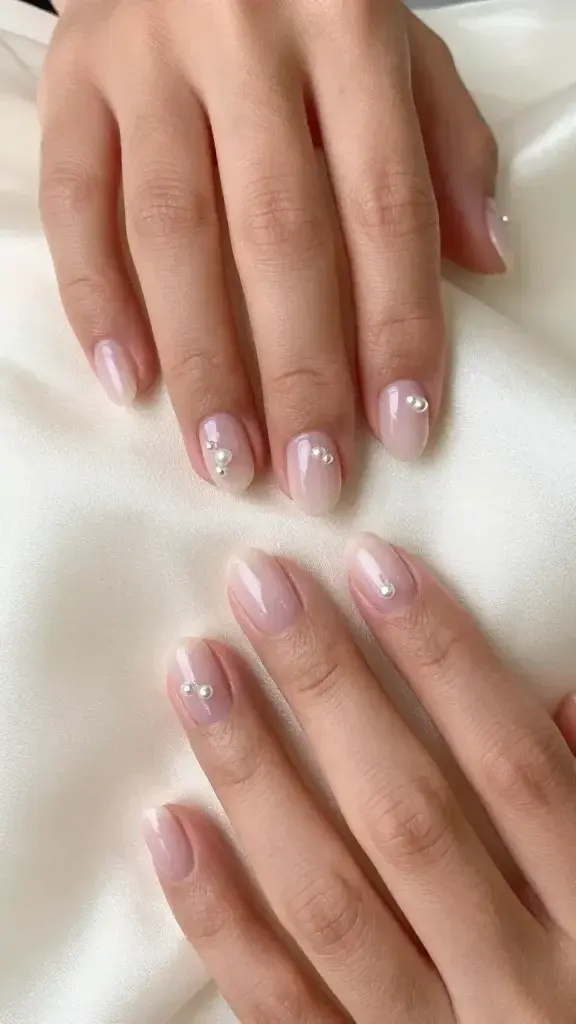 Top-down, hyper-realistic studio photo of both hands resting lightly on a soft ivory silk fabric, showcasing short-to-medium oval nails with a sheer milky pink jelly base (slightly translucent, natural nail line softly blurred). Each nail features 2–3 tiny micro-pearls (1–2 mm), placed asymmetrically near the cuticle or slightly off-center for subtle dimension—no additional gems or glitter. Skin tone is warm medium-tan with natural texture, moisturized cuticles, and a soft, healthy sheen. Lighting is diffused, bright, and natural, creating gentle highlights on the pearls and a glossy but not overly reflective nail finish. Depth of field sharp on nails and pearls, softly blurred background. Color palette: milky blush pink, ivory, warm skin undertones. Minimalist composition, no visible jewelry, clean, modern aesthetic suitable for bridal or date-night inspiration.