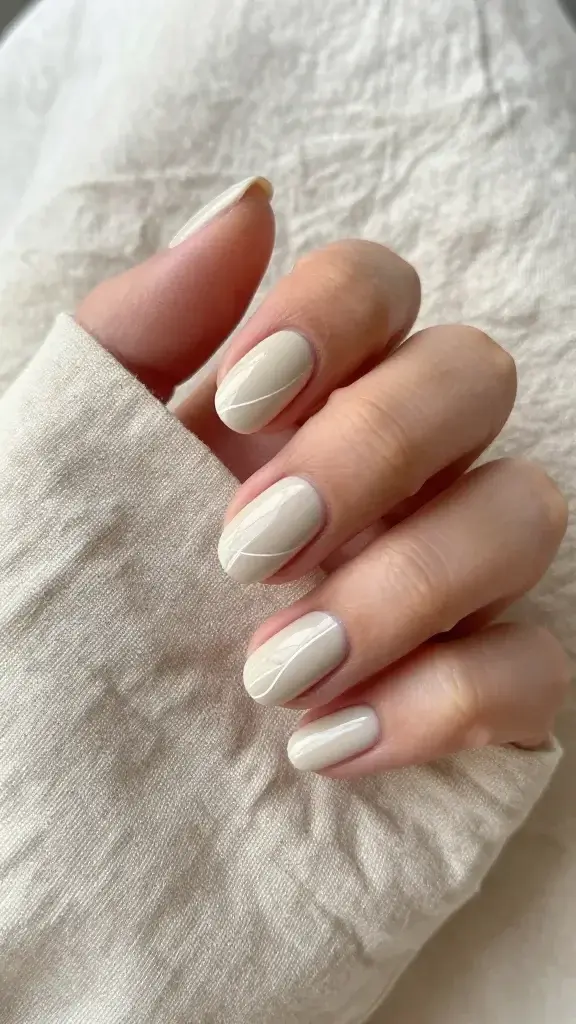 Top-down, hyper-realistic close-up photo of a well-groomed hand resting on softly wrinkled natural linen fabric, showcasing short-to-medium oval nails painted in a creamy linen-beige gel polish with a glossy finish. Each nail features precise, ultra-thin white micro-line designs: index with a single off-center vertical line for length, middle with a diagonal micro-stripe near the tip slanting from outer corner to inner edge, ring with a minimal half-outline tracing the free edge curve, pinky with a subtle diagonal micro-stripe at the tip, and thumb with a clean off-center vertical line. Cuticles are hydrated and neat, skin tone light-to-medium neutral, soft diffused daylight from the left, shallow depth of field emphasizing crisp, architectural linework. No jewelry, no text, no logos—just minimal, modern editorial styling in a beige-on-beige palette.
