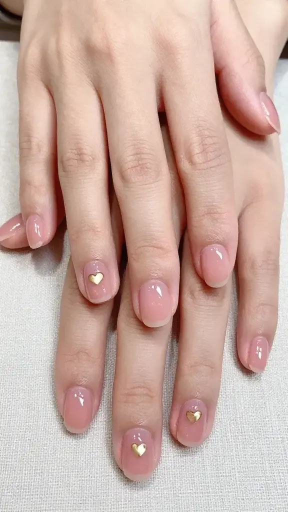Top-down, hyper-realistic studio photo of two well-groomed hands resting on a soft, neutral linen surface, showcasing a minimal short oval manicure with a transparent rosé jelly base (sheer, glossy, dewy finish) on all ten nails. Two accent nails (left ring finger and right index finger) each feature a single tiny metallic gold heart charm, centered just above the cuticle, ultra-thin and micro-sized for a delicate look. The jelly pink is translucent with visible natural nail beds and subtle free-edge glow, no opacity or streaks. Cuticles are hydrated and tidy, skin tone is natural medium-light with warm undertones. Lighting is bright diffused daylight to enhance the glassy shine, with soft reflections and no harsh shadows. No additional art, no glitter, no rhinestones. Include a clear top coat effect that looks fully sealed and snag-free. Composition is symmetrical and close-cropped on the nails, background uncluttered, color palette warm and romantic. Camera: 50mm equivalent, f/5.6, ISO 200, sharp focus on nail surfaces.