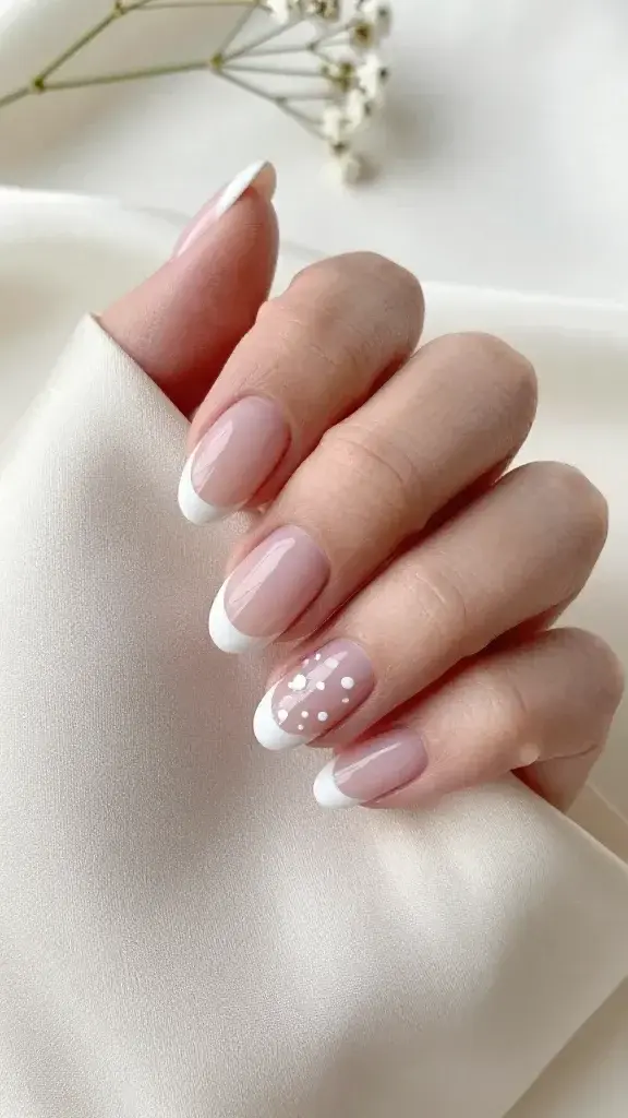 Top-down, high-resolution, realistic photo of a single hand resting on a soft ivory silk fabric, showcasing medium-length almond nails with a whisper-white French tip. Base is a sheer, cool-toned pink that subtly matches fair-neutral skin. The French tips are milky, softened white with a slightly rounded, delicate smile line (not stark or opaque). On two accent nails (ring and middle), add tiny baby’s-breath-inspired micro dots in crisp white, clustered in groups of three and five near the cuticle and subtly trailing toward the mid-nail; keep each dot very petite and varied in size to read floral, not polka dot. Nails are glossy with a glass-like top coat, cuticles clean and hydrated. Lighting is soft and diffused, daylight-like, emphasizing the milky translucence and gentle gradients. Background props minimal (a sprig of real baby’s breath slightly out of focus) to reinforce the airy, romantic, quiet-luxury vibe. Ultra-realistic texture, natural skin tone, no filters, editorial beauty photography style.