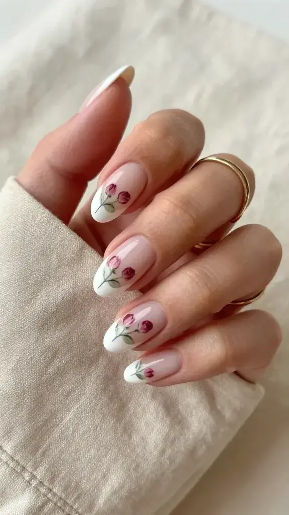 Top-down, hyper-realistic photo of a single hand resting on a soft beige linen surface, showcasing medium-length almond nails with a sheer latte base and crisp, soft-white French tips. Each nail features two to three tiny hand-painted micro-rosebuds placed just above the tip line: dusty rose petals with subtle shading, paired with minuscule sage-green leaves and fine stems, all angled slightly inward toward the nail center for a cohesive, romantic flow. The sheer latte base should allow natural nail beds and a hint of free edge to show through for a “my nails but better” effect. Finish appears ultra-glossy with glass-like reflections, no visible brush strokes. Skin tone warm-neutral with natural texture, moisturized cuticles. Subtle gold stacking rings on the index and ring fingers to emphasize the polished-to-evening versatility. Soft, diffused daylight with gentle shadows; neutral, minimalist composition; extremely high detail macro focus on nail art, bokeh background. Photorealistic, beauty editorial quality.