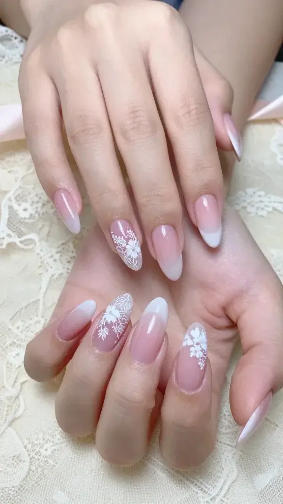 Top-view, high-resolution realistic photo of both hands with almond-shaped nails resting on a soft ivory lace fabric, showcasing a “Vintage Lace Florals Over Pink Jelly” manicure. Nails have a semi-sheer, cool pink jelly base applied in thin, even layers, allowing natural nail beds to subtly show through for depth. Tips feature a diffused French gradient (soft, hazy transition) blending the jelly pink into milky white. Delicate white lace floral motifs are stamped/hand-painted at the tips and outer corners of each nail, with fine filigree, tiny blossoms, and airy negative space that appears to float. Design is consistent but slightly varied per nail for realism. Finish is glossy, glassy shine with soft natural lighting and minimal shadows; skin tone is fair-neutral with hydrated cuticles. Background includes softly out-of-focus lace texture and a hint of a blush silk ribbon for romantic ambiance. Ultra-feminine, modern, soft-glam aesthetic; capture micro details of the lace lines and the translucent jelly depth. 50mm lens, natural daylight, no harsh filters.