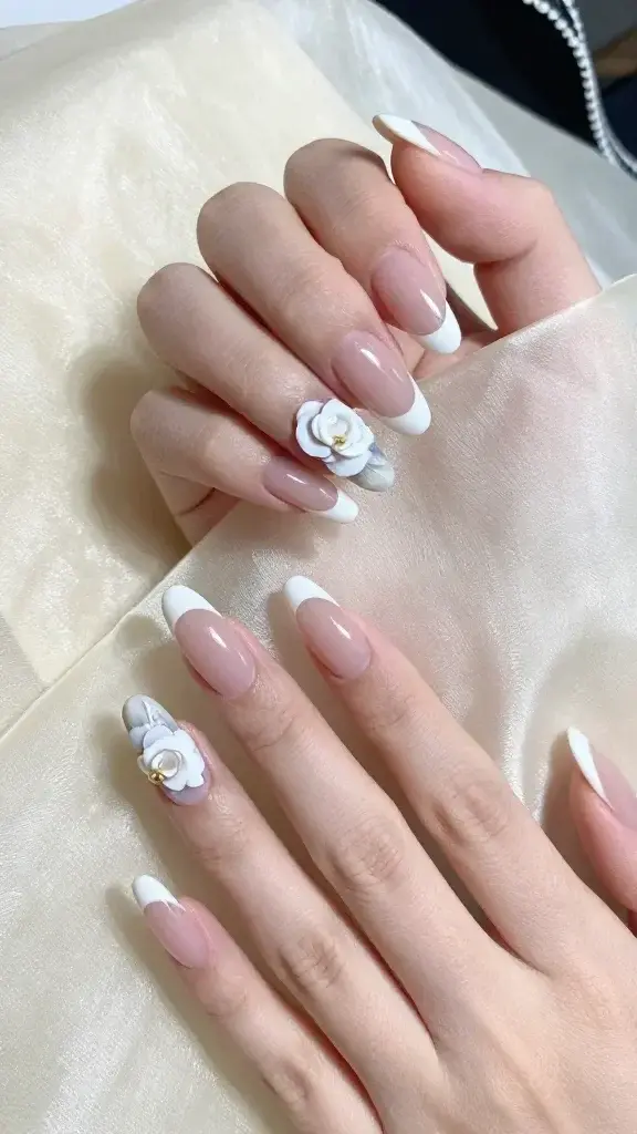 Top-down, hyper-realistic photo of two elegant hands with medium-length almond nails resting on a soft ivory silk fabric, natural daylight from the left creating gentle shadows. Nails feature a classic French manicure: neutral sheer pink base with crisp, thin white tips, high-gloss finish. On each hand, the ring finger is a feature nail with a single hand-painted white camellia: layered soft white petals with subtle cool gray shading for depth, slightly translucent edges, centered near the mid-nail. A tiny gold micro-bead sits at the camellia’s center for a refined accent. Cuticles are immaculate, skin softly moisturized with a natural sheen, no visible hangnails. Color palette: sheer blush pink, chalk white, cool gray shadows, micro gold detail. Background includes a hint of a black tweed ribbon with white trim in the corner for a subtle Chanel-coded vibe, out of focus. Composition is clean, luxurious, editorial beauty lighting with shallow depth of field focusing on the feature nails. No text, no logos, no watermarks.