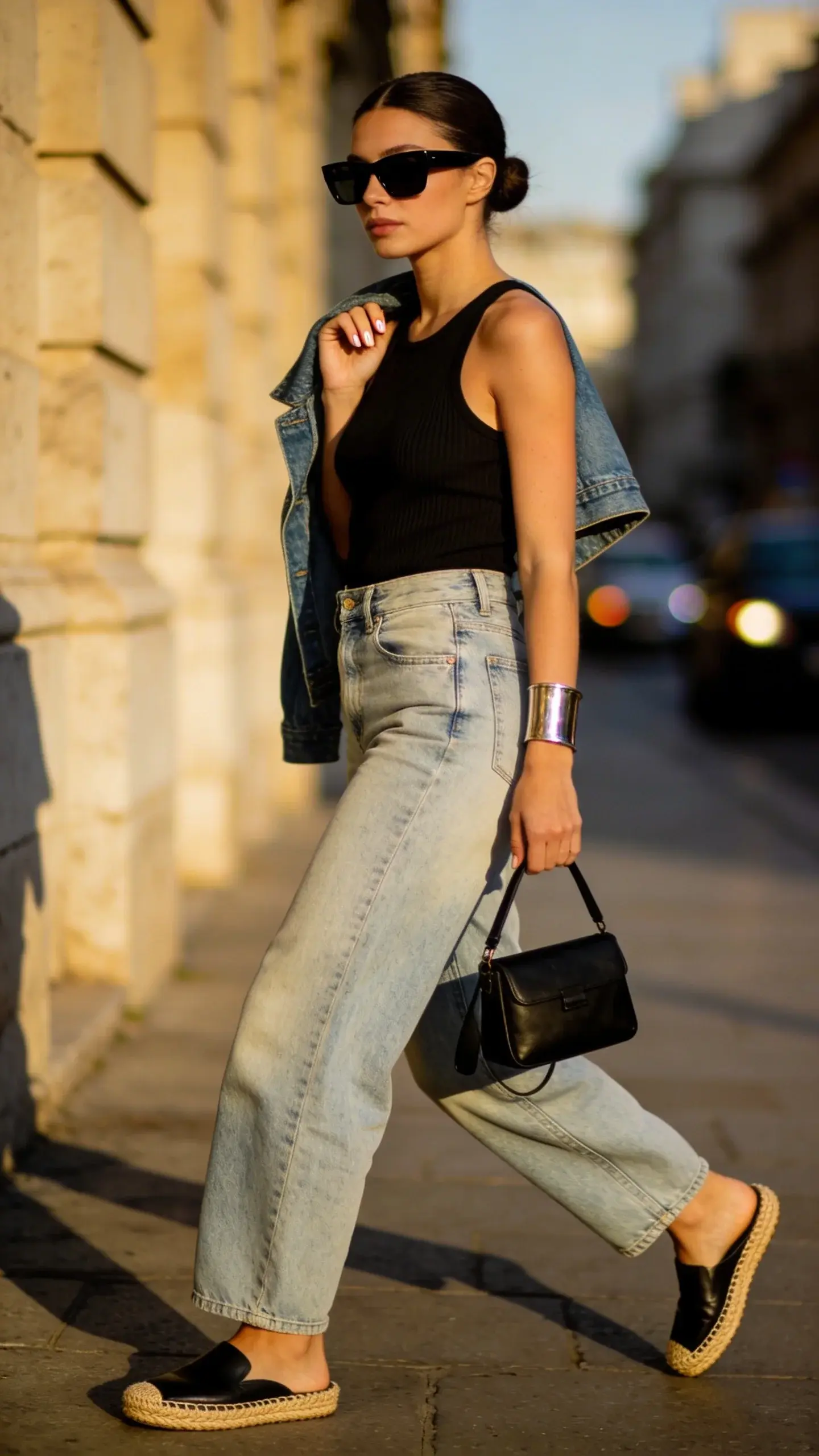 Realistic street-style fashion photo of a chic woman wearing a ribbed fitted tank (black) tucked into high-rise barrel-leg jeans in a light vintage wash, paired with low-profile black leather espadrilles (flat, no ankle ties). She accessorizes with a single polished metal cuff bracelet, a structured mini shoulder bag in black with a short strap under her arm, and matte black rectangular sunglasses. Add a subtle cropped denim jacket draped over her shoulders for layering. Setting: golden-hour city sidewalk in a stylish urban neighborhood (neutral stone buildings, soft shadows, bokeh traffic). Composition: three-quarter body, walking mid-stride, confident posture, natural candid expression. Lighting: soft, warm, directional sunlight highlighting textures (ribbed knit, denim grain, rope espadrille edge). Styling details: clean center-part low bun, minimal makeup, short natural nails. Color palette: monochrome and denim blue with metal accents. Emphasize modern proportions—fitted top balancing voluminous curved-leg jeans—while keeping the espadrilles sleek and understated. Ultra high-resolution, editorial fashion photography, shallow depth of field, crisp detail, no text.