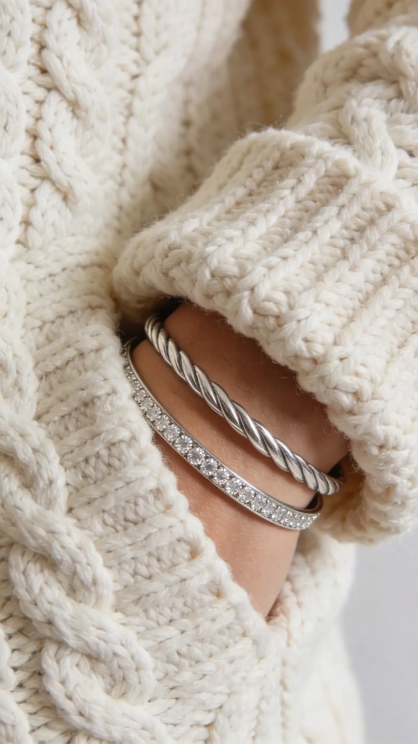 Fashion close-up of layered textures: cuff of a chunky cream cable-knit sweater pushed back to reveal two slim cable bangles and one pavé diamond station bracelet; soft diffused light, tactile emphasis on knit and twisted metal, cozy elevated style, no text.