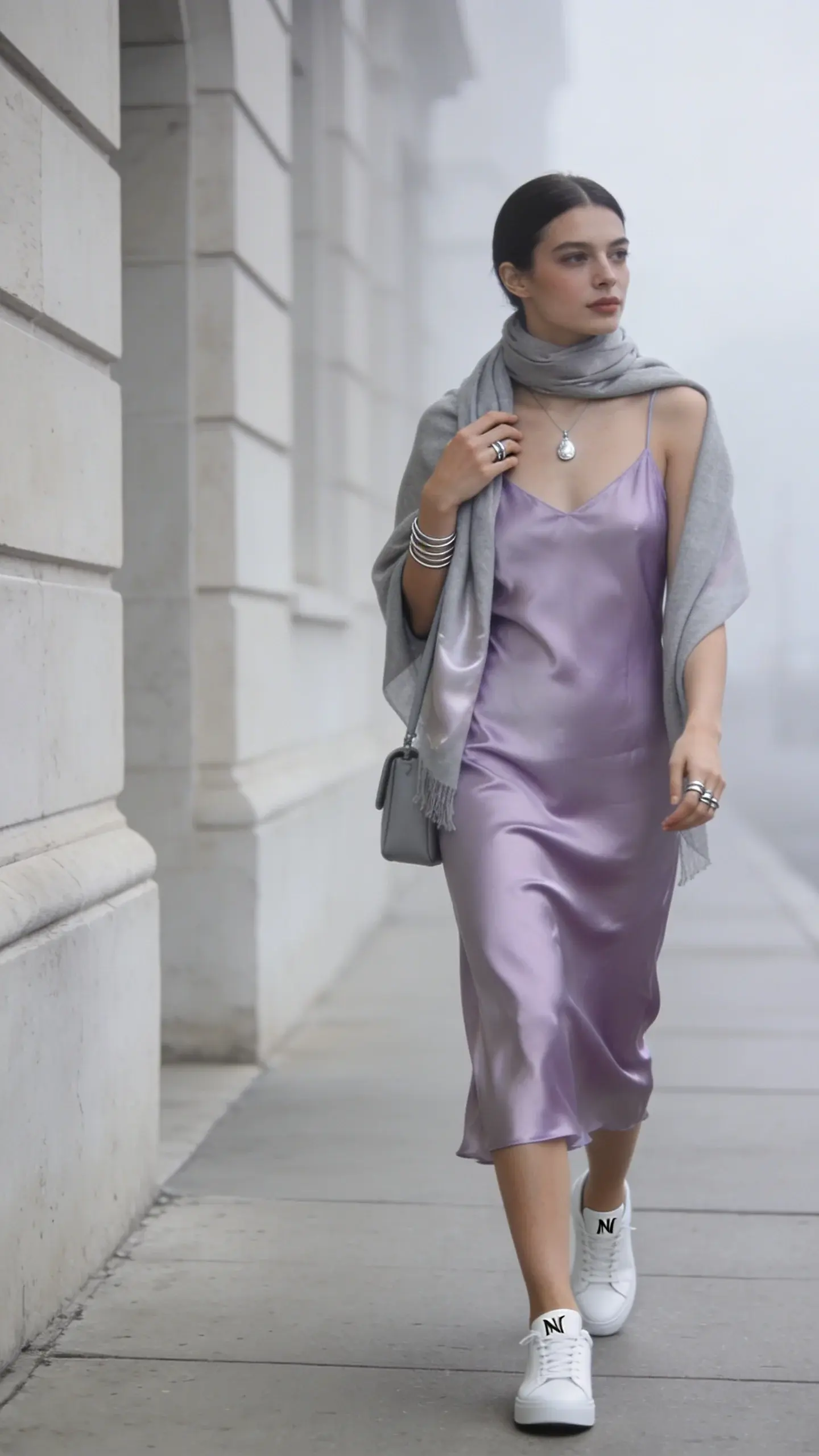 Realistic high-fashion street style photo of a woman embodying the “Cold Summer” color palette and soft luxury aesthetic, walking along a minimalist city sidewalk near pale stone architecture in soft overcast daylight. She wears a misty lavender satin midi slip dress in a cool periwinkle-lavender tone, bias-cut so it skims the body with a subtle sheen. On her feet: pristine white low-profile leather sneakers with minimal branding. Draped lightly around her neck and shoulders: a summer-weight light gray cashmere-silk scarf. Accessories: a delicate silver pendant necklace, thin silver stacking rings, and a soft slate mini shoulder bag with a short strap tucked under her arm. Styling is effortless and polished: clean, luminous skin, natural makeup with cool undertones, soft straight hair tucked behind one ear. She moves with relaxed confidence, mid-stride, one hand grazing the scarf. Color grading emphasizes cool, misty tones (lavender, slate, gray, white) with gentle contrast and fine fabric texture detail. Shallow depth of field, 50mm lens look, high-resolution editorial photography, no logos or text.