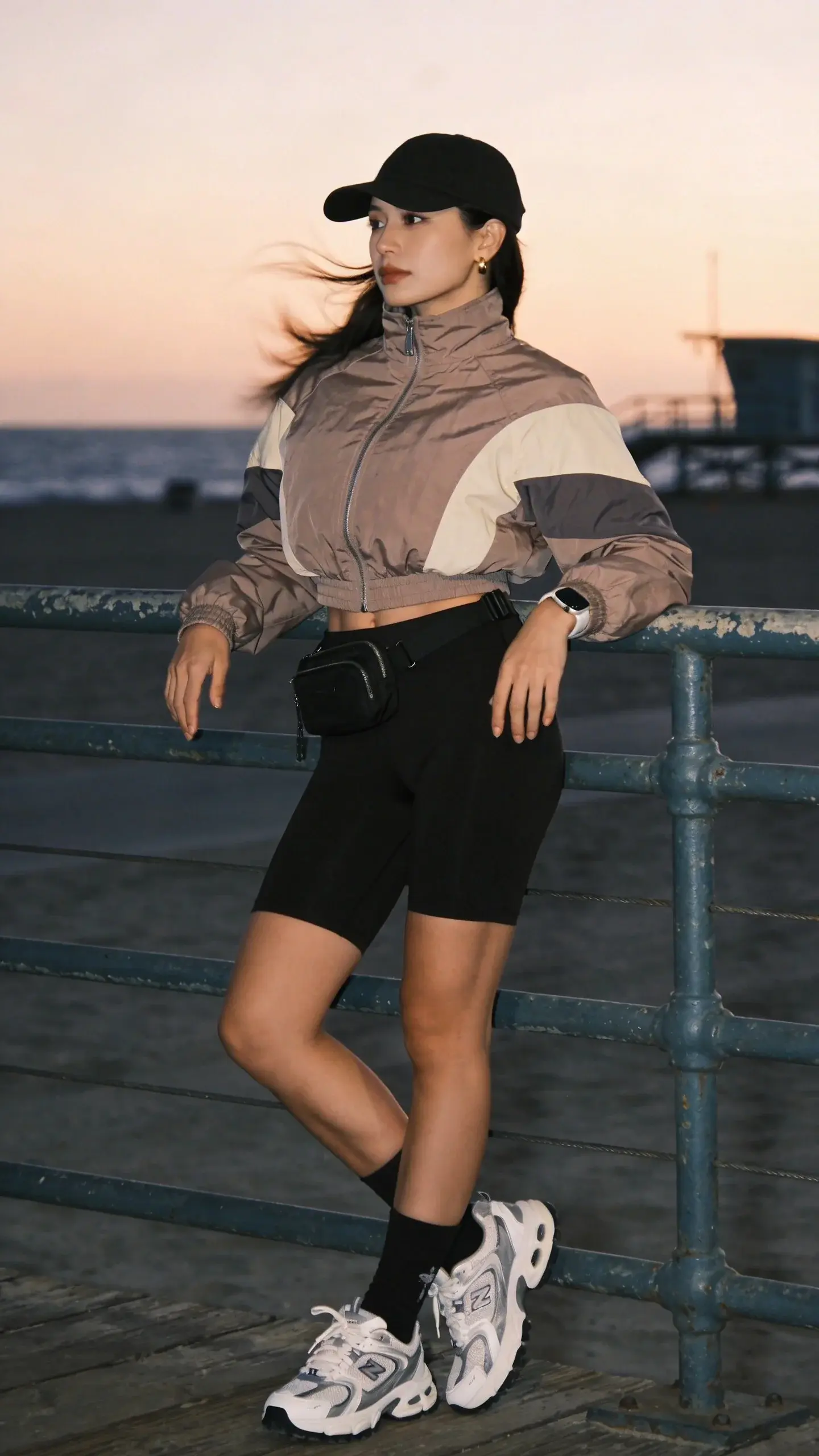 Golden-hour beach boardwalk scene featuring a chic woman dressed for a cold summer night: she wears a sleek, lightweight windbreaker (neutral taupe base with subtle color-block panels in ivory and charcoal), zipped halfway with a slightly cropped fit, paired with high-rise black bike shorts that hit mid-thigh. On her feet are retro white-and-gray chunky trainers with cushioned soles and ankle socks. She tops the look with a matte black baseball cap and minimal gold stud earrings, plus a black mini belt bag worn crossbody and a slim sporty watch. She casually leans against a weathered railing, ocean breeze catching the windbreaker for a sense of motion; background shows a softly blurred shoreline, pastel sunset sky, and a hint of lifeguard tower. Lighting is warm and cinematic with cool undertones from the sea air, crisp details on technical fabric textures and sneaker treads. Realistic fashion photography, full-body portrait, shallow depth of field, 35mm lens look, no text, editorial street-style vibe.