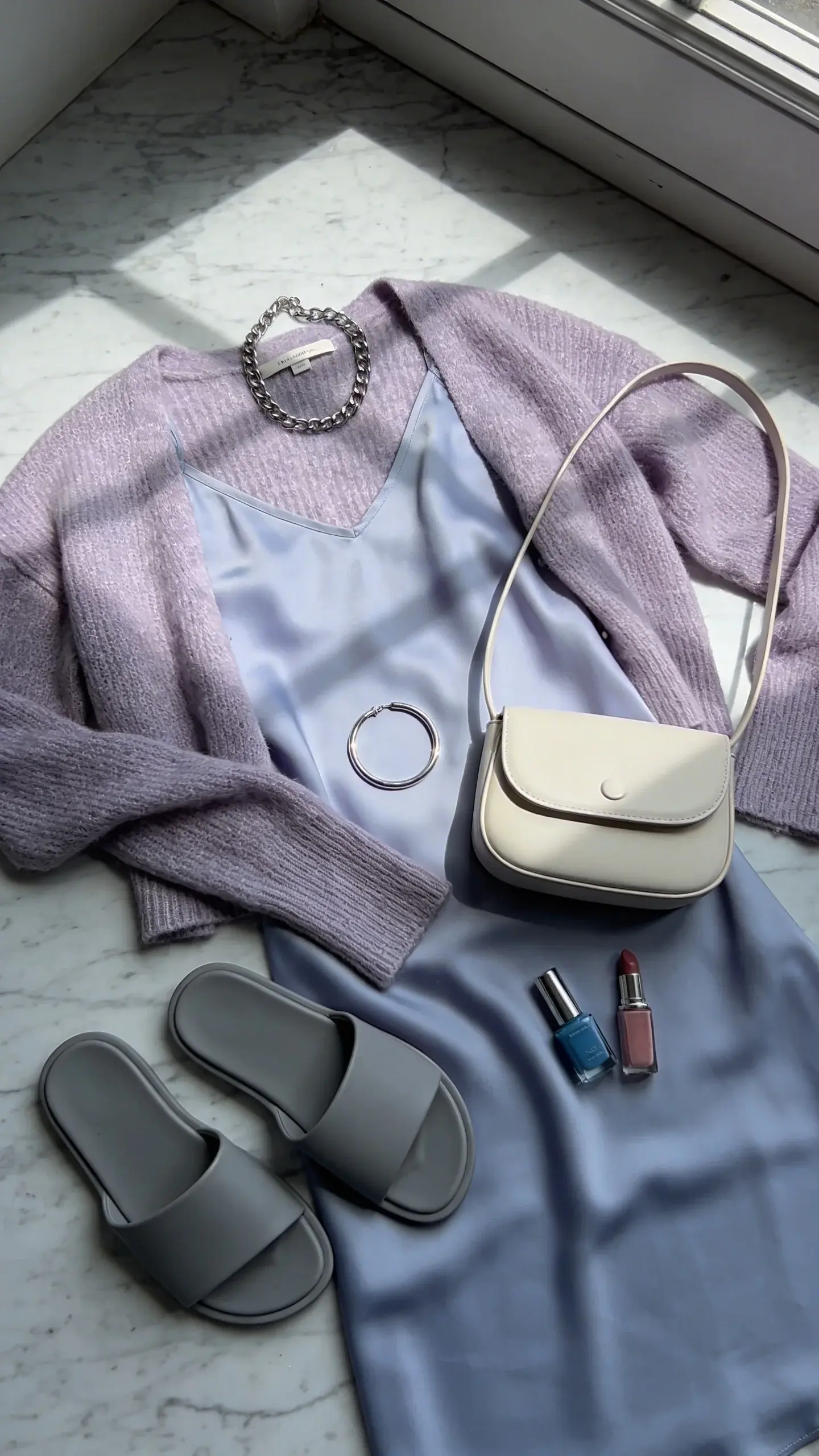 Close-up fashion flat lay on a marble surface: periwinkle silk slip dress, frosted-lilac lightweight cardigan draped beside it, slate-grey minimalist slides, silver chain necklace and slim silver bangle, bone-colored mini shoulder bag; palette includes powder blue nail polish and cool blush lipstick; soft natural window light with gentle shadows; refined, cool-toned styling; no text.
