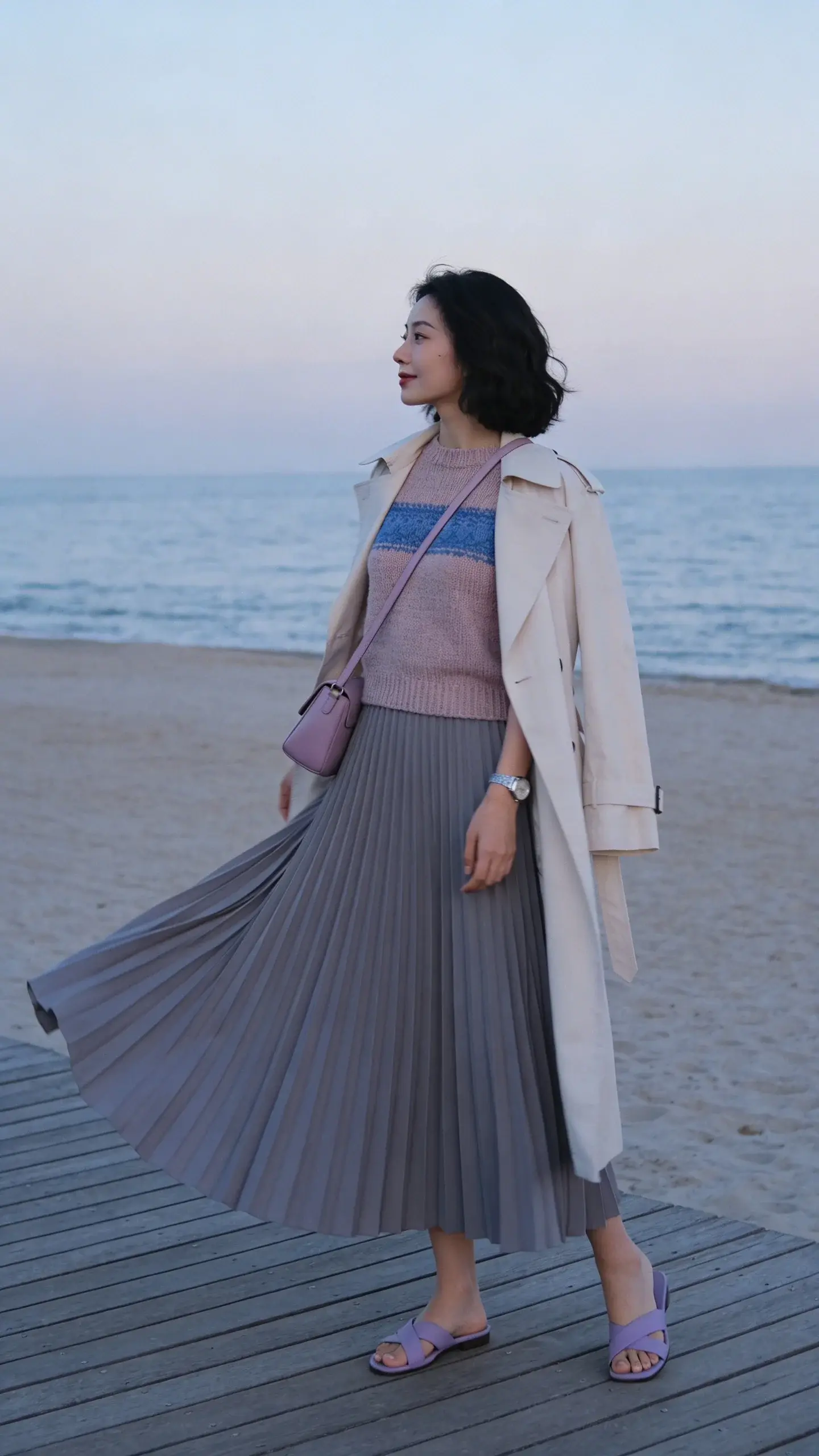 Beach boardwalk evening look: woman in a long flowing dove-grey pleated skirt with a fitted cool-blush (blue-undertone) knit top, layered with a light bone trench draped over shoulders; periwinkle crossbody bag, silver minimalist watch, soft lavender sandals; wind catching the skirt, muted ocean and pale sky in background; overall cool, serene palette and elegant silhouette; no text.