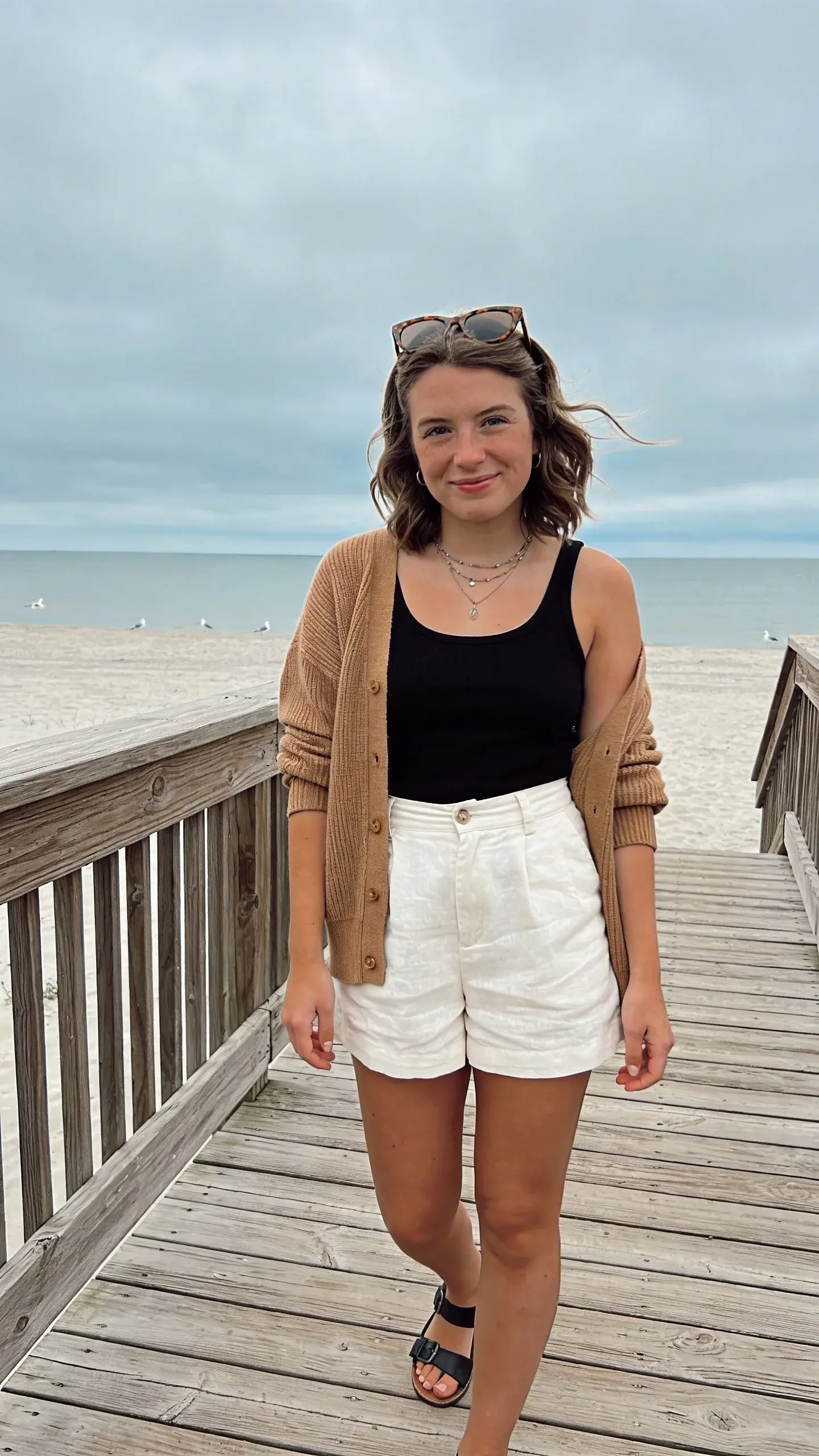 Overcast beach boardwalk with a woman layered in a fine-knit camel cardigan draped over a fitted black tank and high-waisted white linen shorts. Cardigan unbuttoned, sleeves pushed to mid-forearm; black leather thong sandals; tortoiseshell sunglasses on her head; delicate layered necklaces. Hair: shoulder-length waves. Include a light wind, textured wooden planks, muted ocean and gray-blue sky, seagulls in the distance. Color palette: warm neutrals and soft blues. No text.