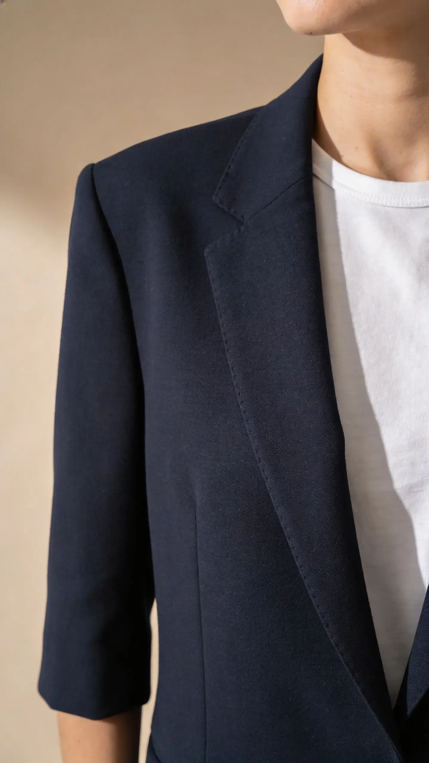 On-body fit checklist visual: cropped torso shot focusing on blazer fit—navy wool-blend blazer over a simple white tee; shoulder seams align perfectly with the model’s shoulders, sleeves ending at the wrist bone, blazer skimming the body without pulling at the button; neutral beige background, subtle side lighting to emphasize structure and drape; clean, modern aesthetic, no text overlays.