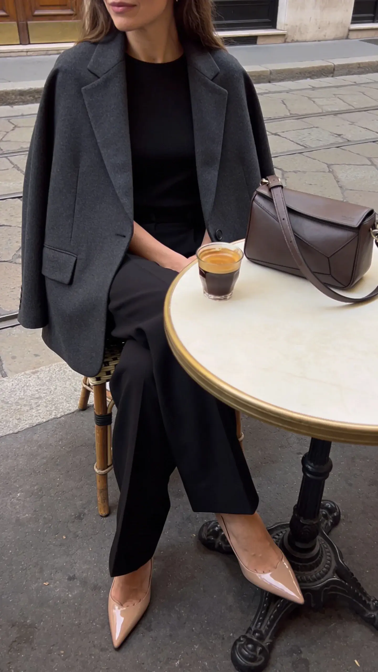 Street style vignette: seated café moment with an elegant woman draping a charcoal blazer over her shoulders (cape style) paired with tailored black trousers and pointed-toe nude pumps; minimalist leather crossbody bag on the table next to an espresso; blazer features minimal pockets, clean lapel line, mid-weight wool that holds shape; soft afternoon light, European sidewalk café setting with muted tones, no text.