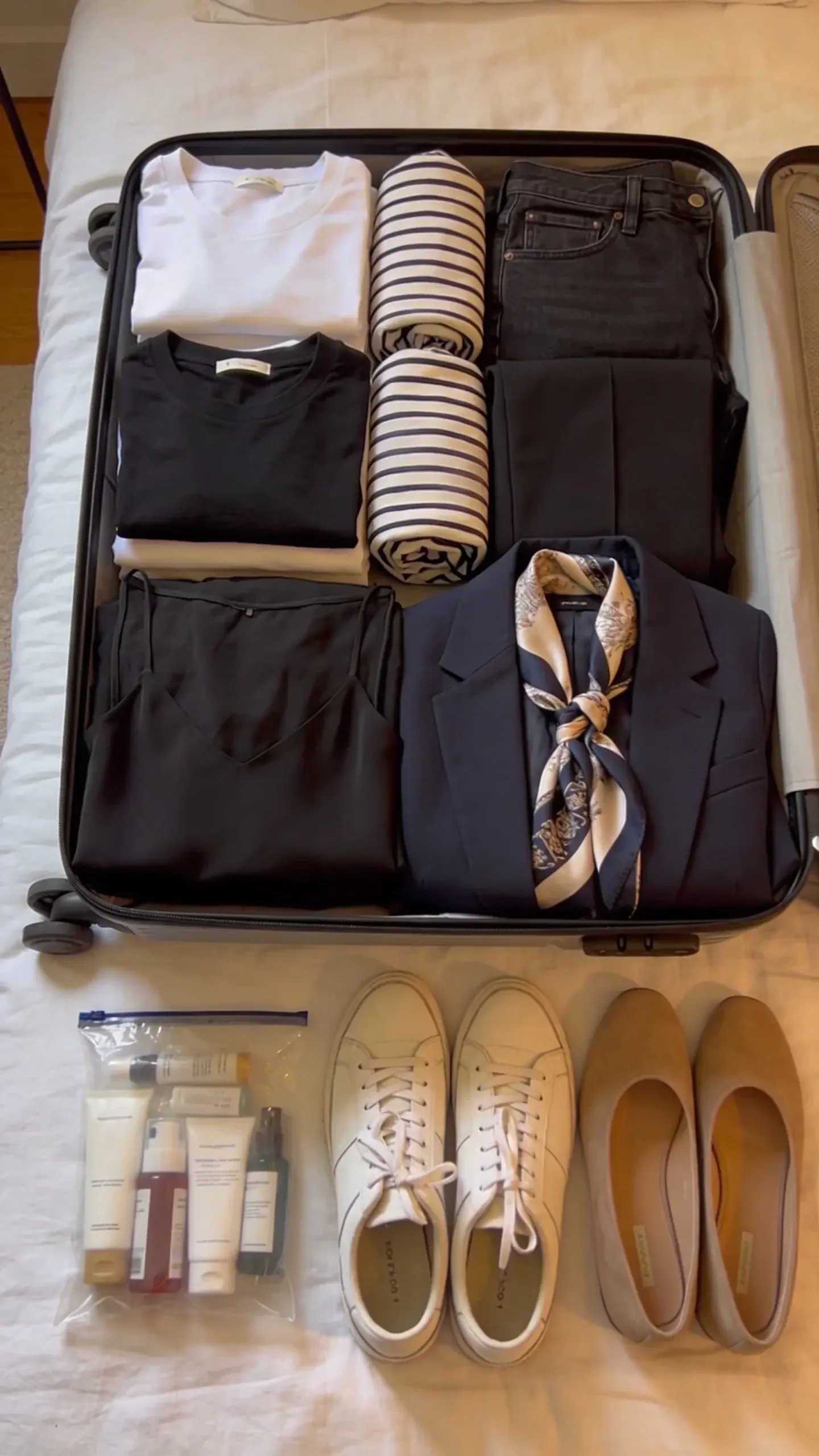A compact capsule wardrobe packing scene: open carry-on suitcase on a bed with neatly rolled basics (two white tees, one black tee, striped long-sleeve, dark jeans, black trousers), folded black slip dress, lightweight navy blazer, silk scarf, minimalist sneakers, loafers placed beside, and travel-sized toiletries in a clear pouch; warm indoor lighting, tidy and intentional layout.