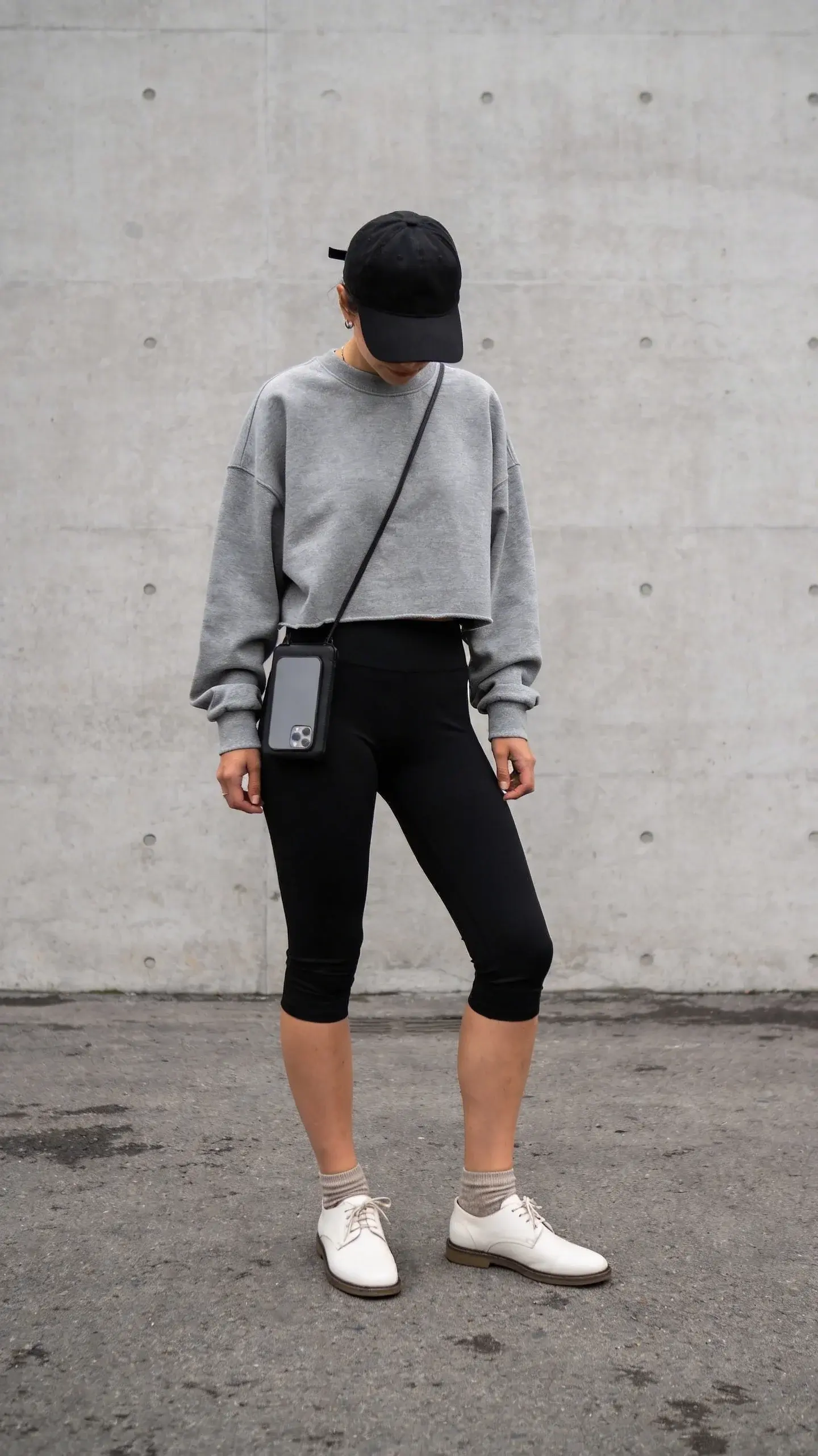 Coffee-run aesthetic: black capris with a gray boxy cropped sweatshirt, white minimalist leather sneakers, ankle socks barely visible, black baseball cap, slim crossbody phone pouch; matte concrete wall background, overcast lighting, no text.