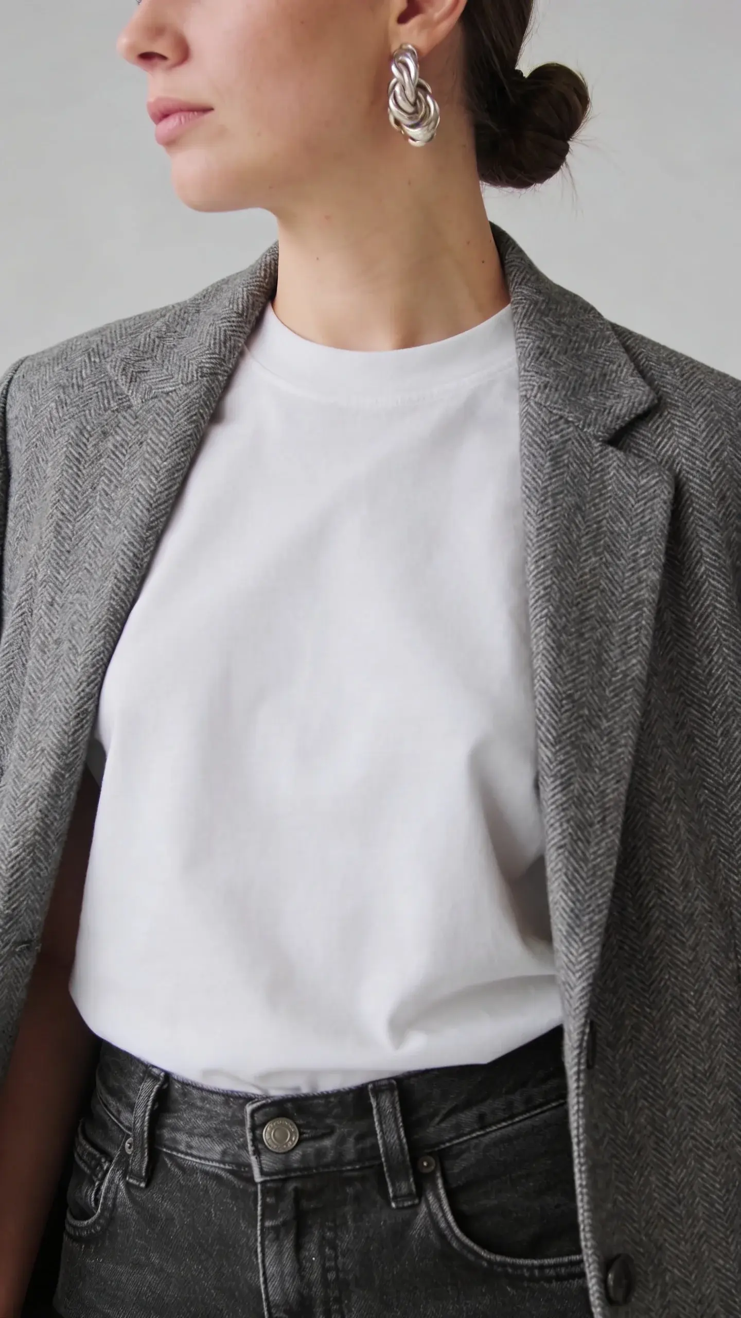 Minimal studio portrait: close-up from waist up of a woman wearing a gray herringbone blazer draped open over a bright white cotton-modal tee; straight dark-wash jeans visible at the waistband; hair in a low bun, chunky silver statement earrings; clean light-gray backdrop, soft diffused lighting, no text.