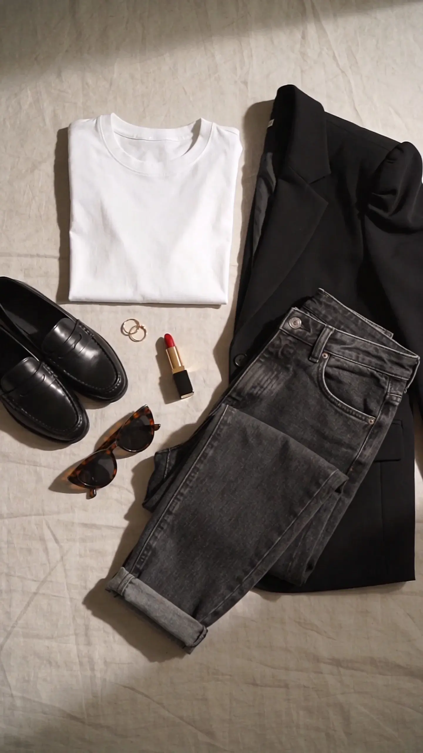 Detail-focused flat lay: neatly arranged crisp white cotton tee, structured black blazer with strong shoulders, straight-leg medium-wash jeans folded to show ankle length; accessories include black leather loafers, tortoiseshell sunglasses, red lipstick tube, small gold hoops; neutral linen background, soft top-down light, no text.