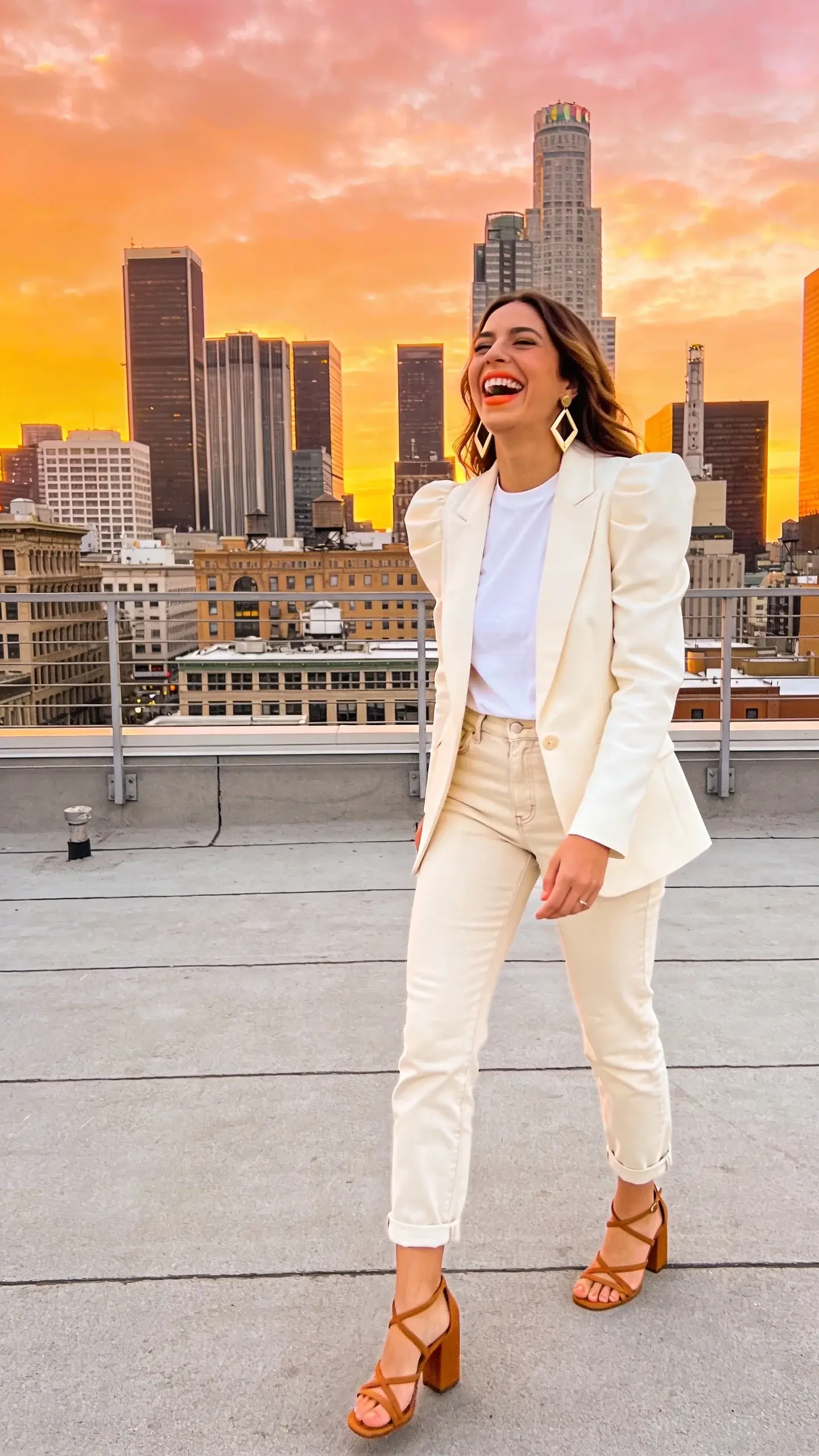 Rooftop sunset gathering: a woman laughing with skyline behind her, ivory blazer with pronounced shoulder line over a bright white tee, light ecru straight jeans cropped at ankle; tan strappy block-heel sandals, coral lip, statement geometric earrings; warm sunset glow, candid composition, no text.