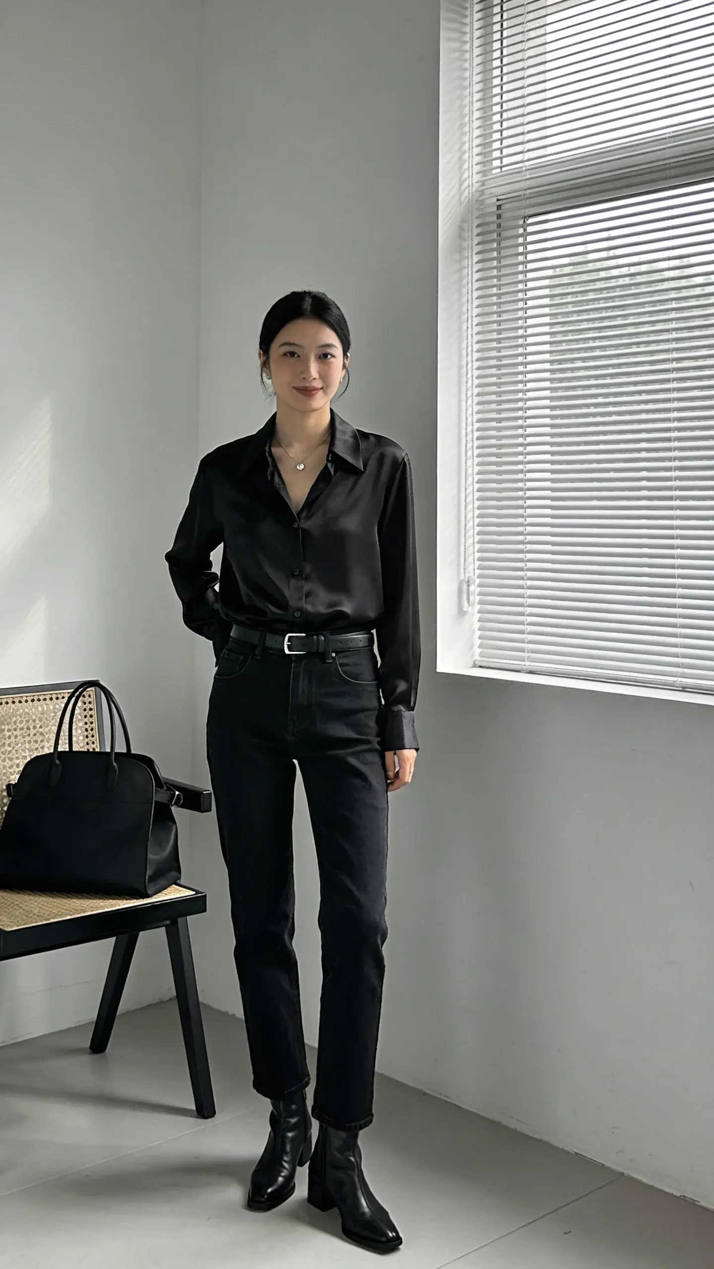 Sleek all-black ensemble in a minimalist apartment: person in a black silk button-down tucked into matte black straight-leg jeans, paired with black leather ankle boots; black belt with a small silver buckle; black structured tote on a nearby chair; a delicate silver necklace; window light catching the silk sheen and denim texture; muted grayscale interior.