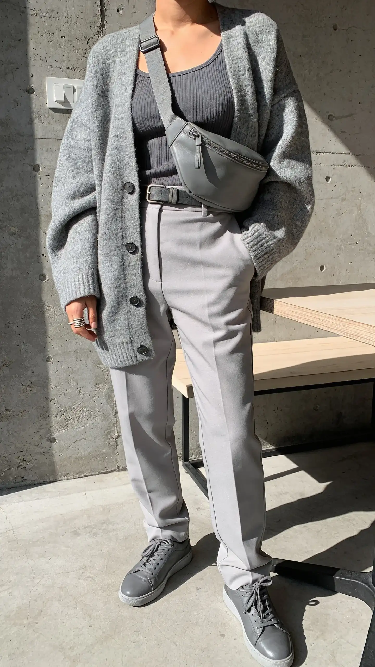 Cool gray monochrome with mixed textures in a modern café: person in a heather-gray oversized cardigan over a charcoal-gray ribbed tank, paired with light-gray tailored trousers; gray leather sneakers; medium-gray belt bag worn crossbody; thin silver stacking rings; concrete wall and pale wood table for a minimalist backdrop; warm afternoon light.