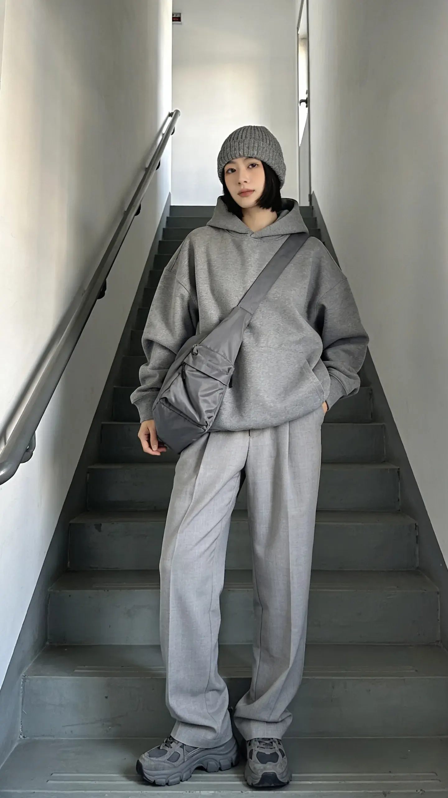 Soft gray athleisure-chic on a staircase: person in a dove-gray oversized hoodie, light-gray tailored joggers, and gray knit beanie; slate-gray chunky sneakers; medium-gray nylon crossbody bag; stairwell with white walls and clean lines; morning light emphasizing cozy-to-sleek texture mix.