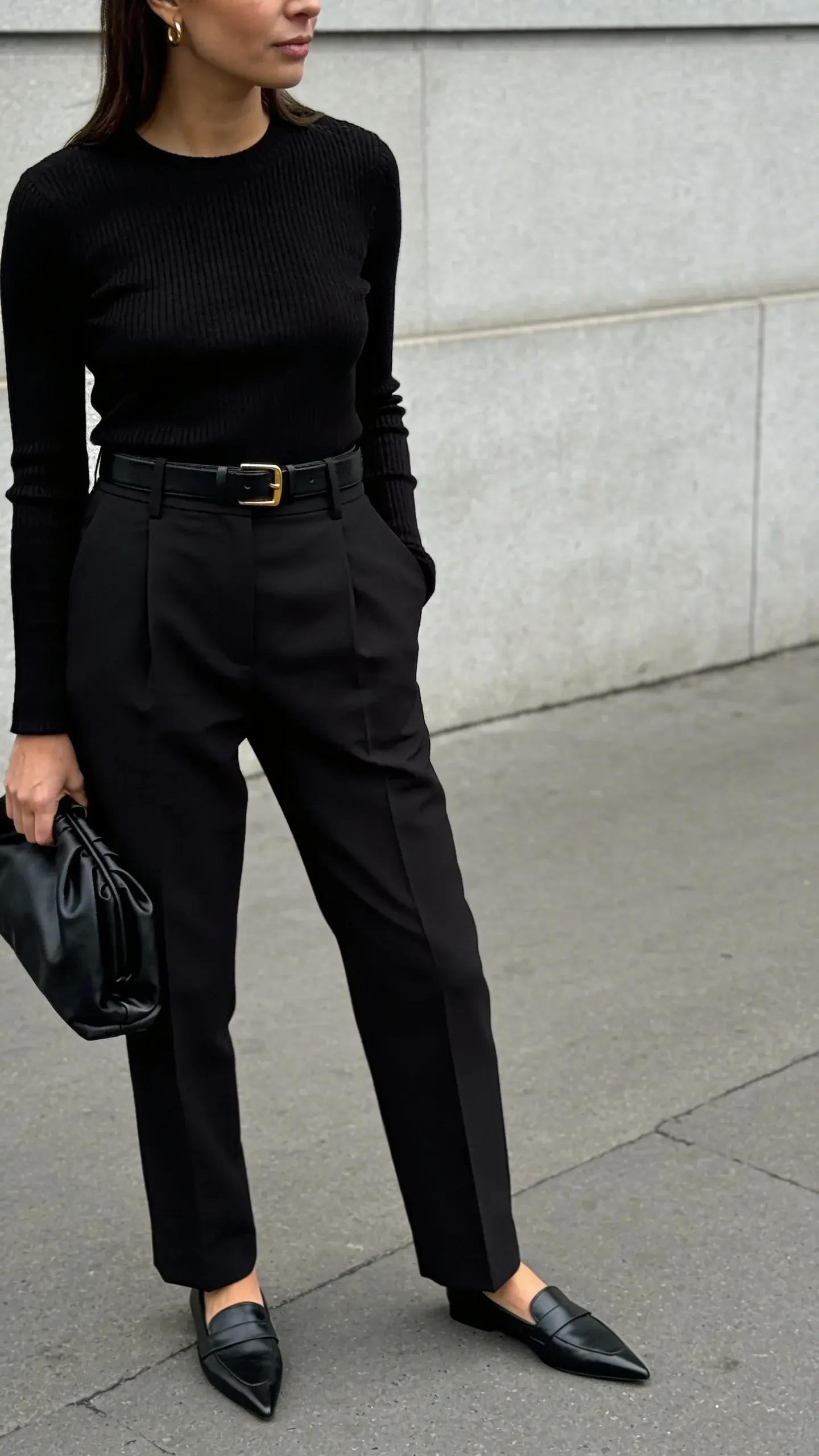 A chic street-style shot of a woman in an all-black monochrome outfit: fine-gauge black knit top, high-waisted tailored black trousers with sharp crease, black leather belt with minimal gold buckle, black pointed-toe loafers, small gold hoop earrings; subtle texture mix with a satin black clutch; photographed in soft overcast light against a light gray urban wall; no text.
