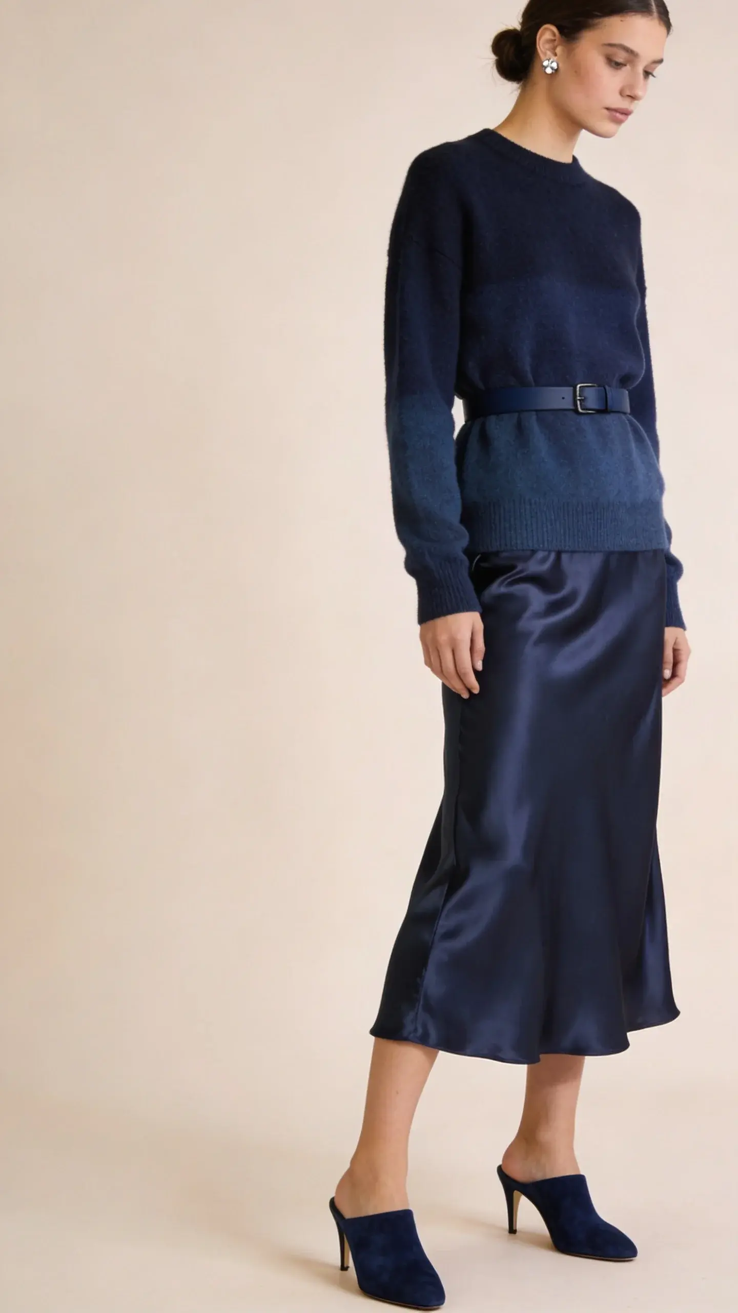 A minimalist studio portrait of a model wearing head-to-toe navy: silk satin slip skirt and merino crewneck sweater in slightly different navies, navy suede kitten-heel mules, slim navy belt, delicate silver studs; hair in a low bun; clean backdrop in pale beige; focus on fabric sheen contrast.