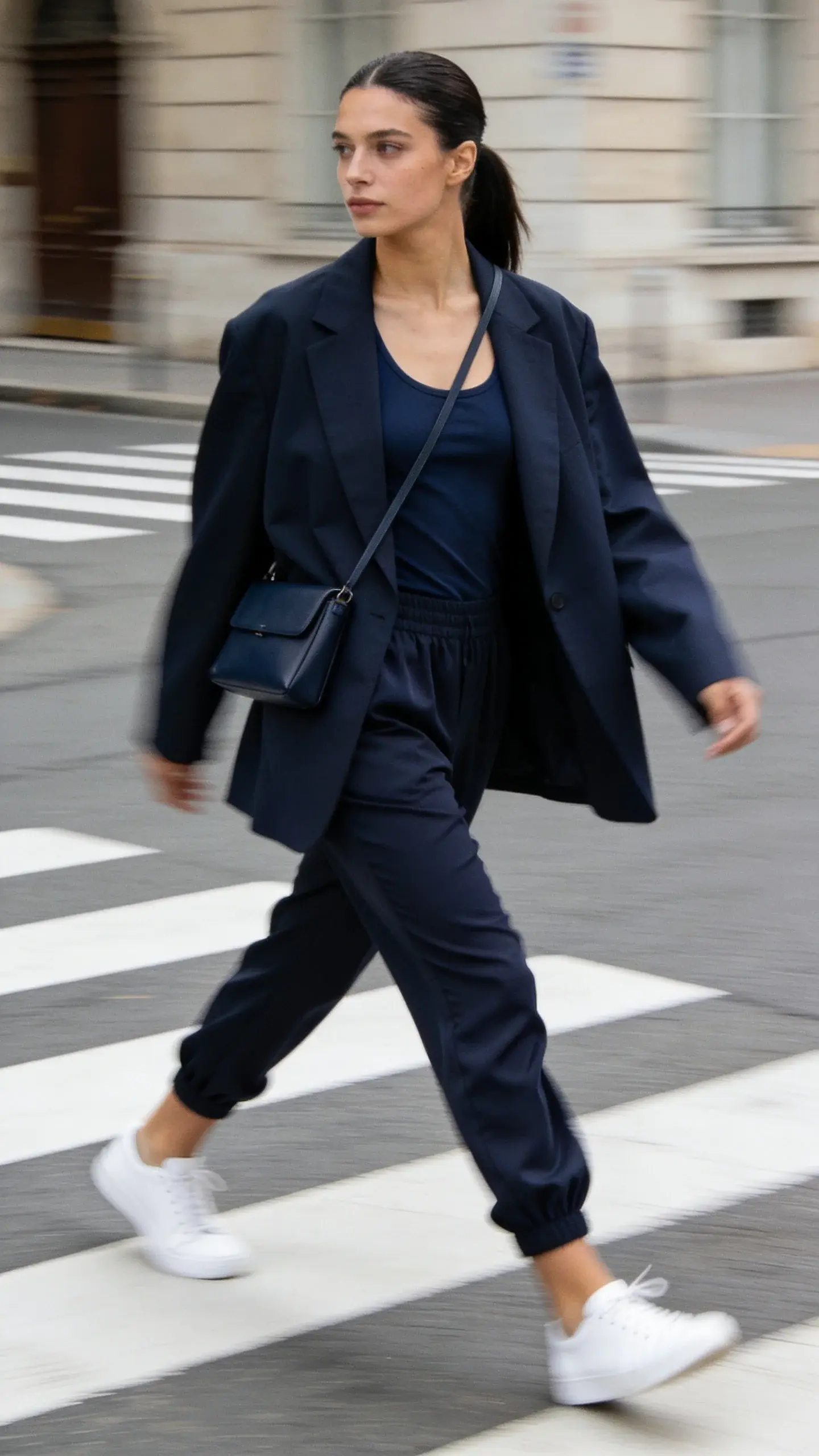 Street-style navy set with sporty polish: navy oversized blazer over navy tank, navy tapered jogger-style trousers in crepe, white minimalist sneakers, slim navy crossbody bag, sleek ponytail; shot mid-stride on a crosswalk; clean lines, dynamic movement.