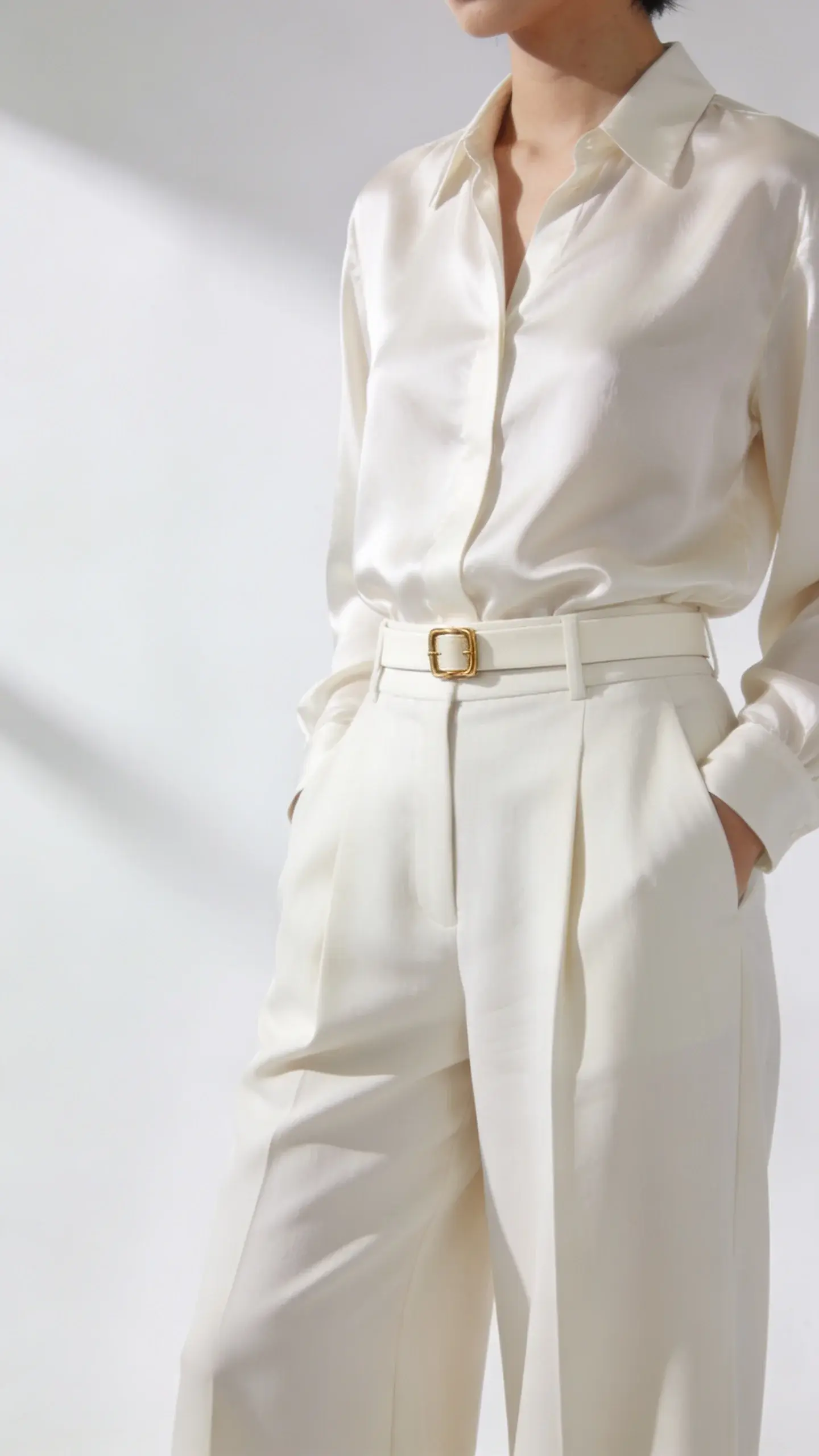 Minimalist monochrome: cream high-waisted wide-leg trousers with a tucked-in cream silk blouse, waist emphasized with a slim 1-inch cream belt with a small gold buckle; bright studio backdrop, soft shadows, clean lines, no text.