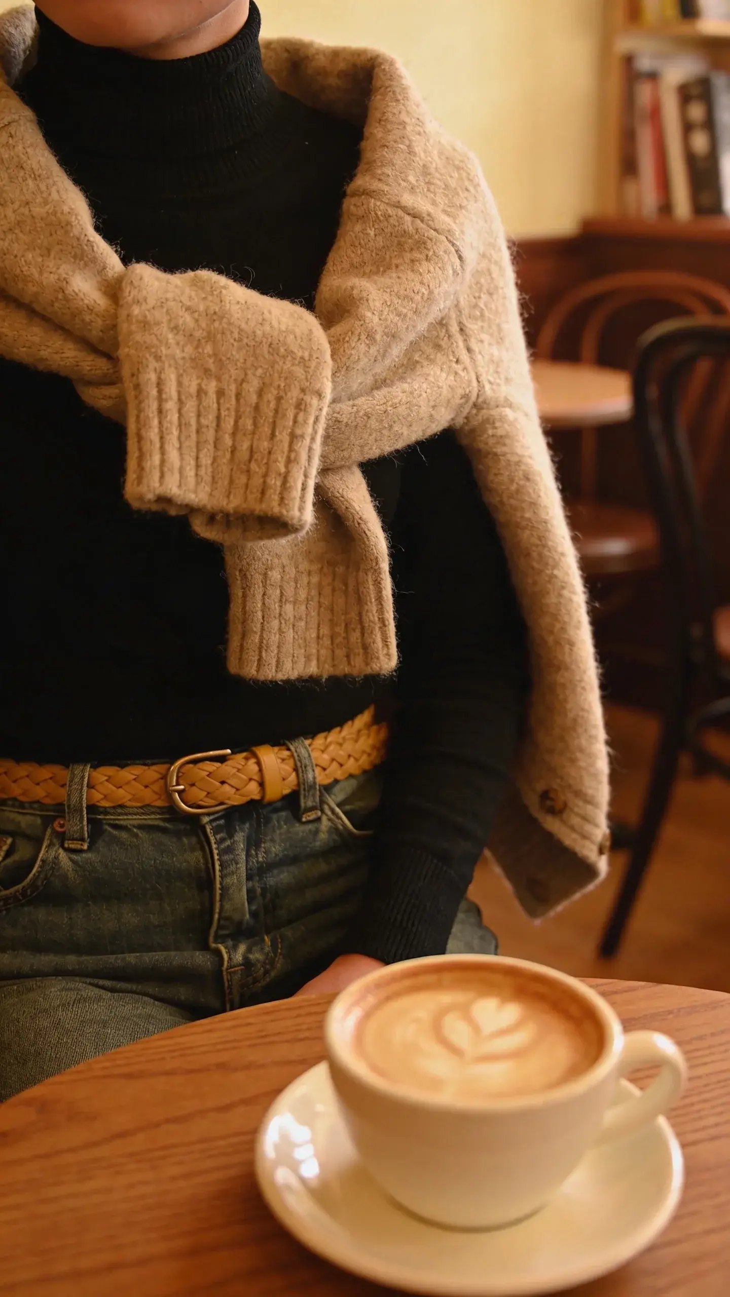 Casual weekend: oversized chunky oatmeal cardigan draped over a fitted black turtleneck and high-rise jeans, waist cinched with a tan braided belt; cozy café corner with a latte on the table, warm ambient light, no text.
