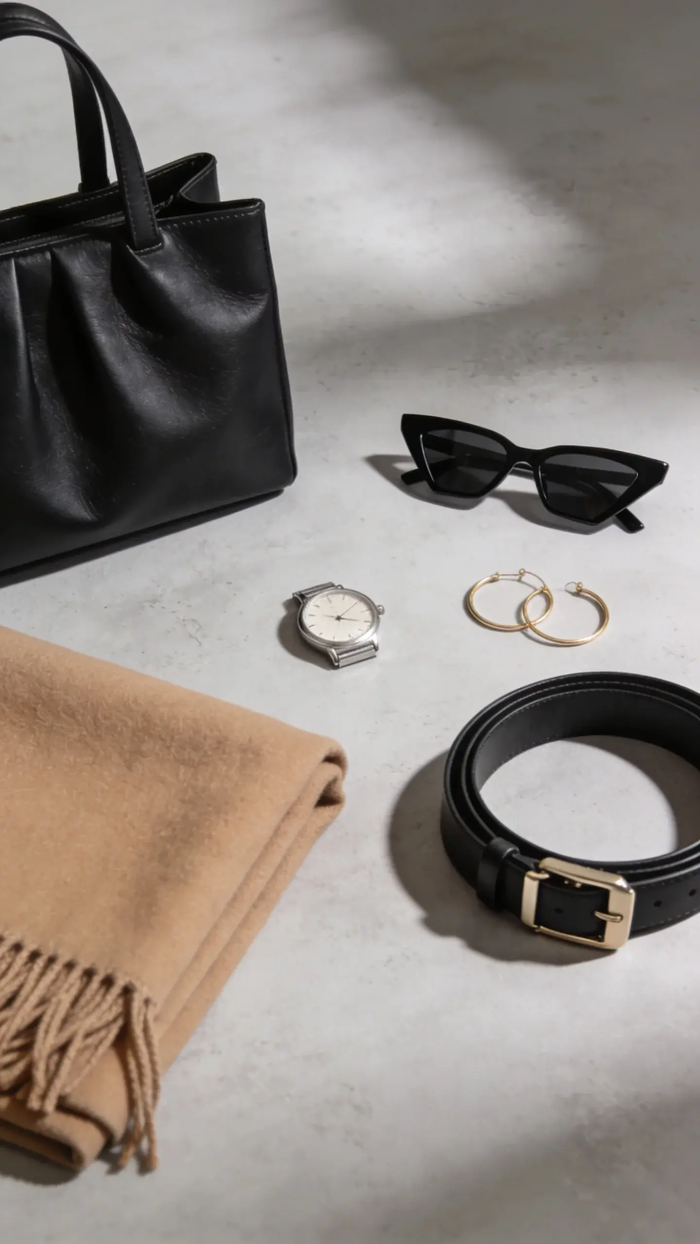 Overhead flat lay of “quiet luxury” accessories: black leather structured tote, slim black sunglasses with angular frames, silver minimalist watch, simple gold hoop earrings, black leather belt with understated buckle, and a neatly folded camel cashmere scarf; set on a textured light gray surface with soft shadows.