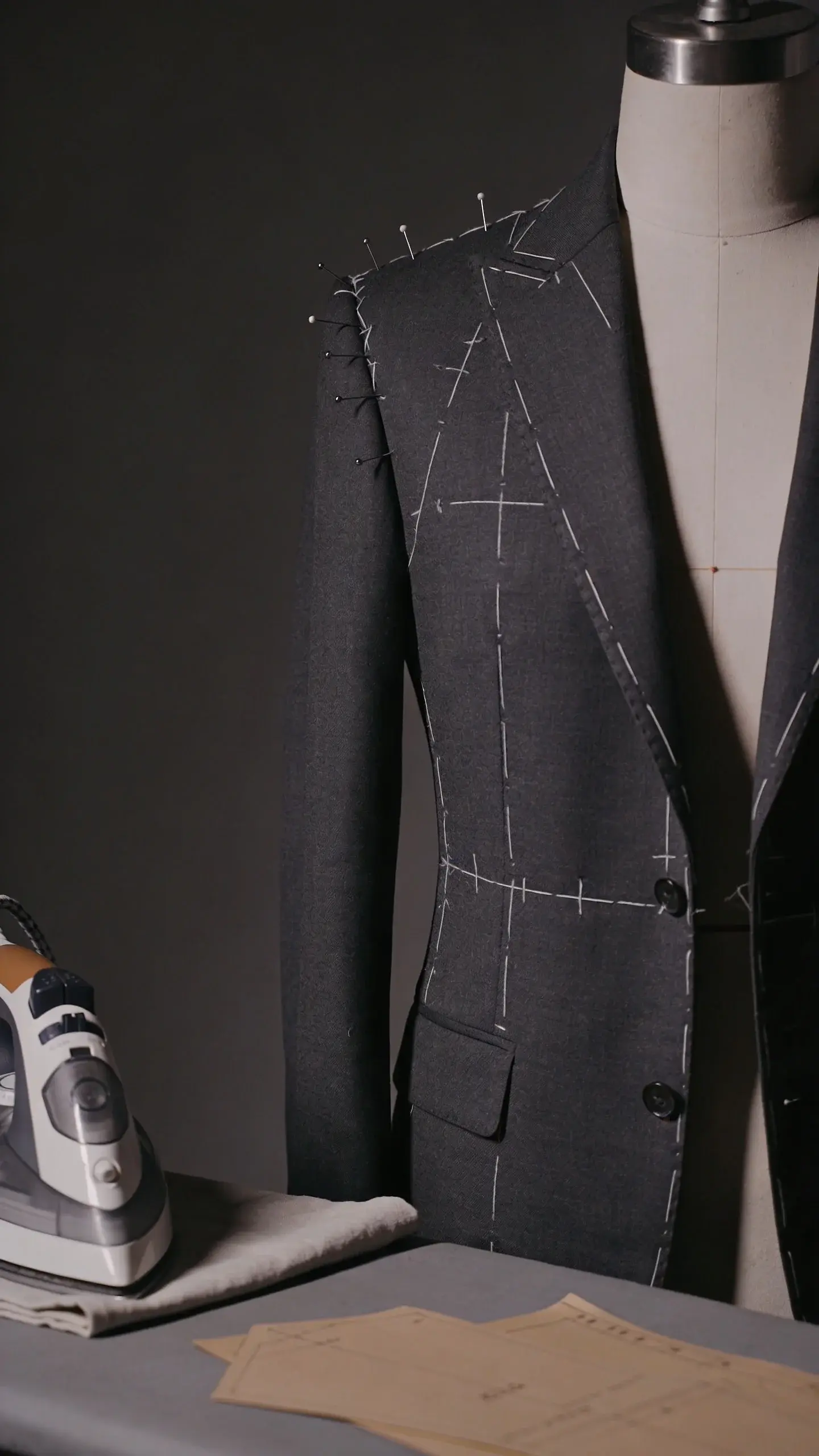 Tailor’s studio scene: a dress form with pins marking shoulder and waist adjustments on a charcoal blazer, chalk lines visible; iron, pressing cloth, and pattern paper nearby; moody, focused light.