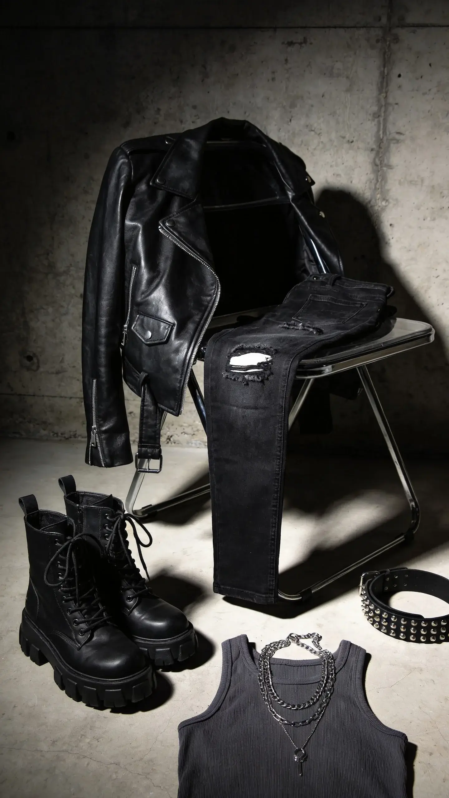 Edgy style personality: A moody urban backdrop with a black leather biker jacket draped over a metal chair, distressed black skinny jeans folded on the seat, chunky lace-up combat boots on the floor, a charcoal ribbed tank, layered silver chain necklaces, and a studded black belt; gritty concrete wall, directional side lighting, high contrast.