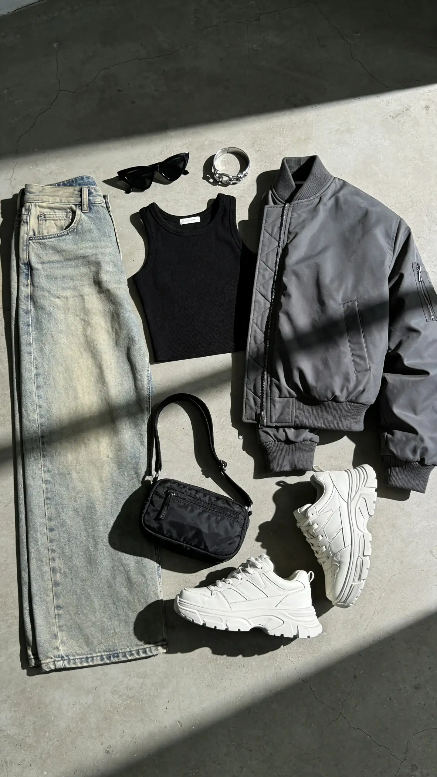 Modern/Street style personality: A concrete loft floor flat lay with oversized light-wash baggy jeans, a cropped black tank, a boxy gray bomber jacket, white chunky sneakers with thick soles, angular black sunglasses, a silver cuff bracelet, and a mini nylon shoulder bag; cool-toned lighting, graphic shadows.