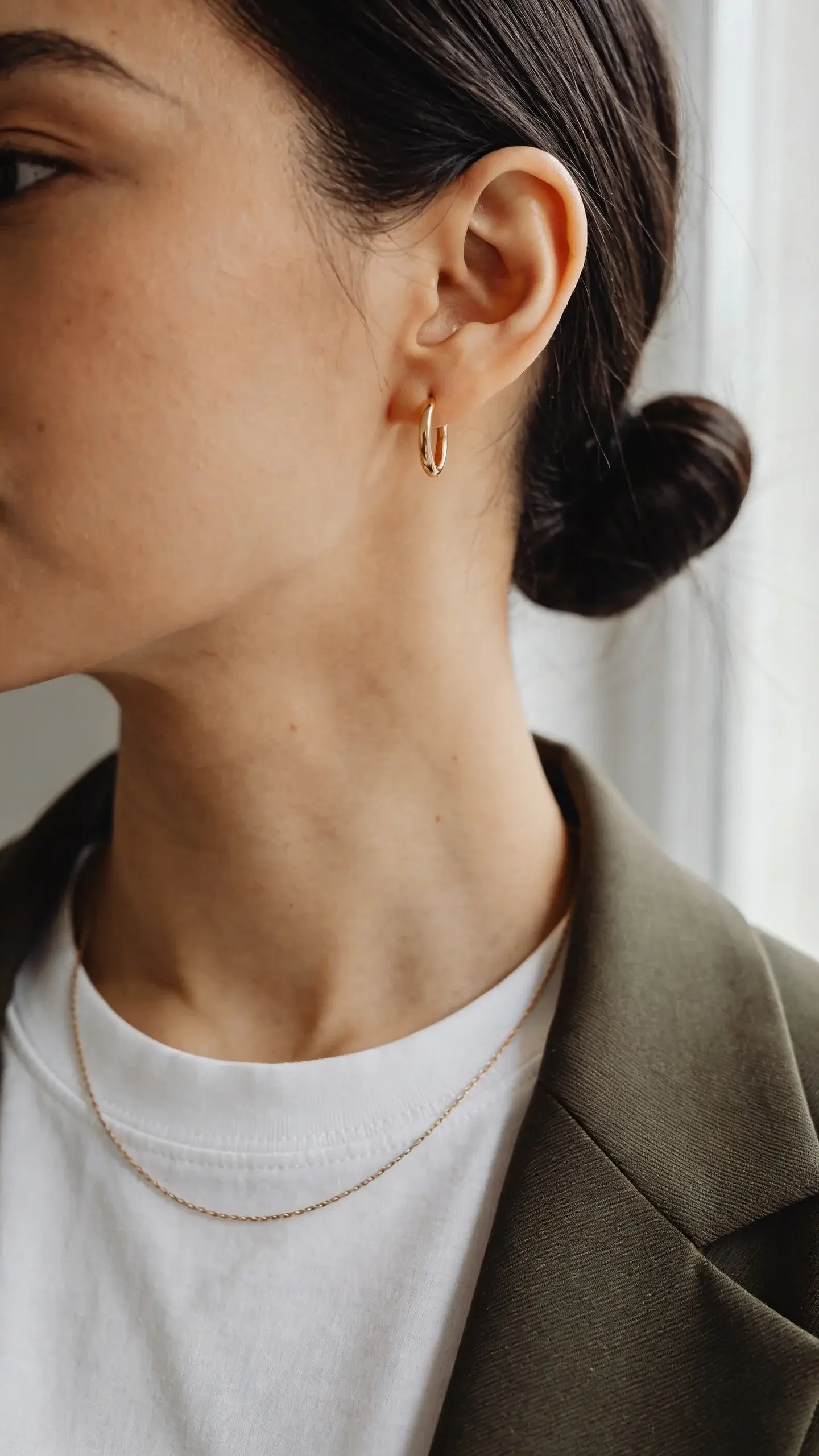 Close-up portrait crop: jawline to chest; smooth low bun, small gold hoop earring, delicate thin chain resting on a clean white crewneck tee, blazer lapel in frame; soft daylight from a window, neutral background, no text.