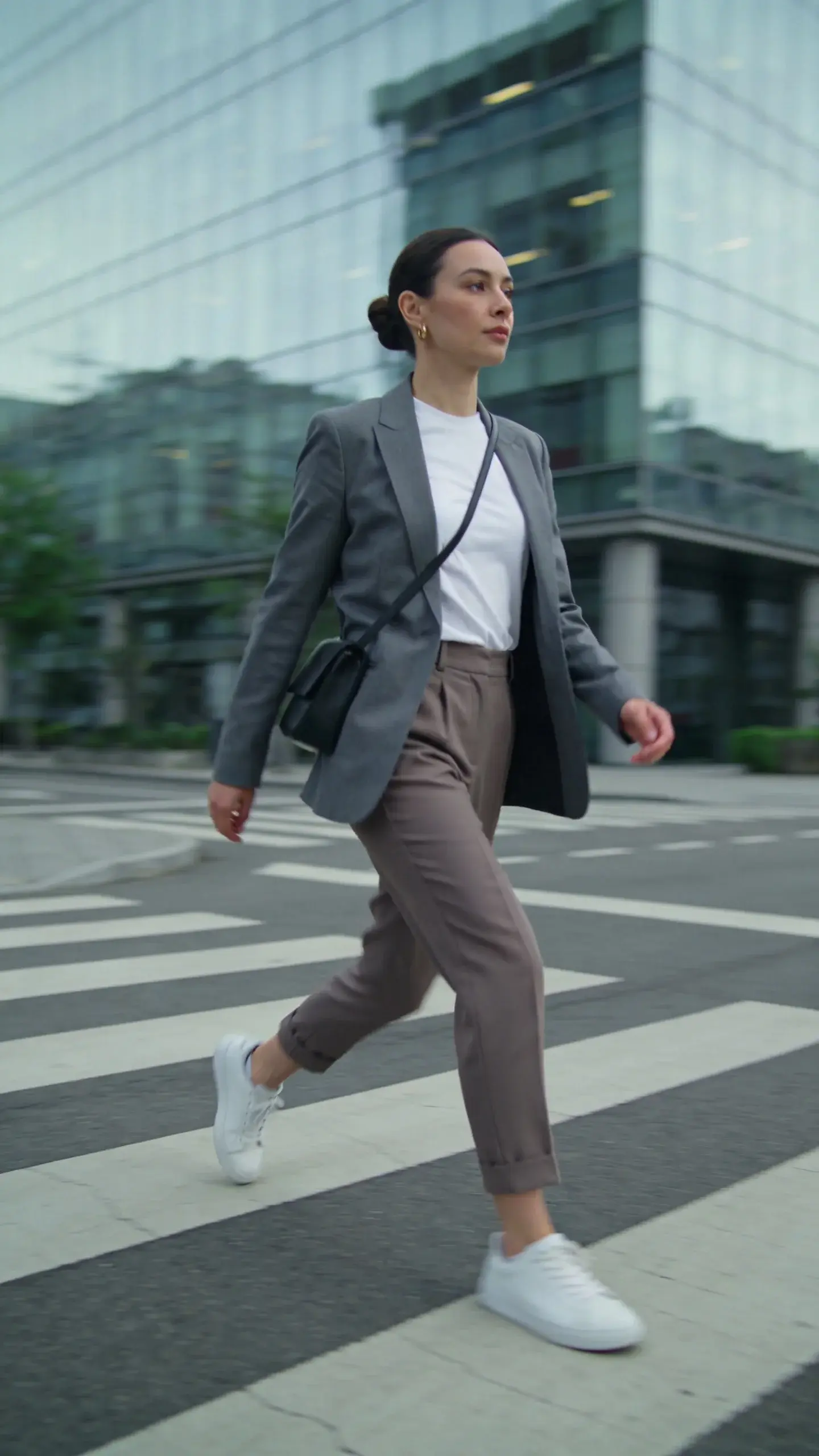 City crosswalk shot: woman mid-stride in taupe tailored ankle trousers, bright white tee, slate gray blazer, crisp white sneakers, slim black crossbody bag; hair in sleek bun, subtle gold earrings; modern glass buildings in background, desaturated color palette, no text.