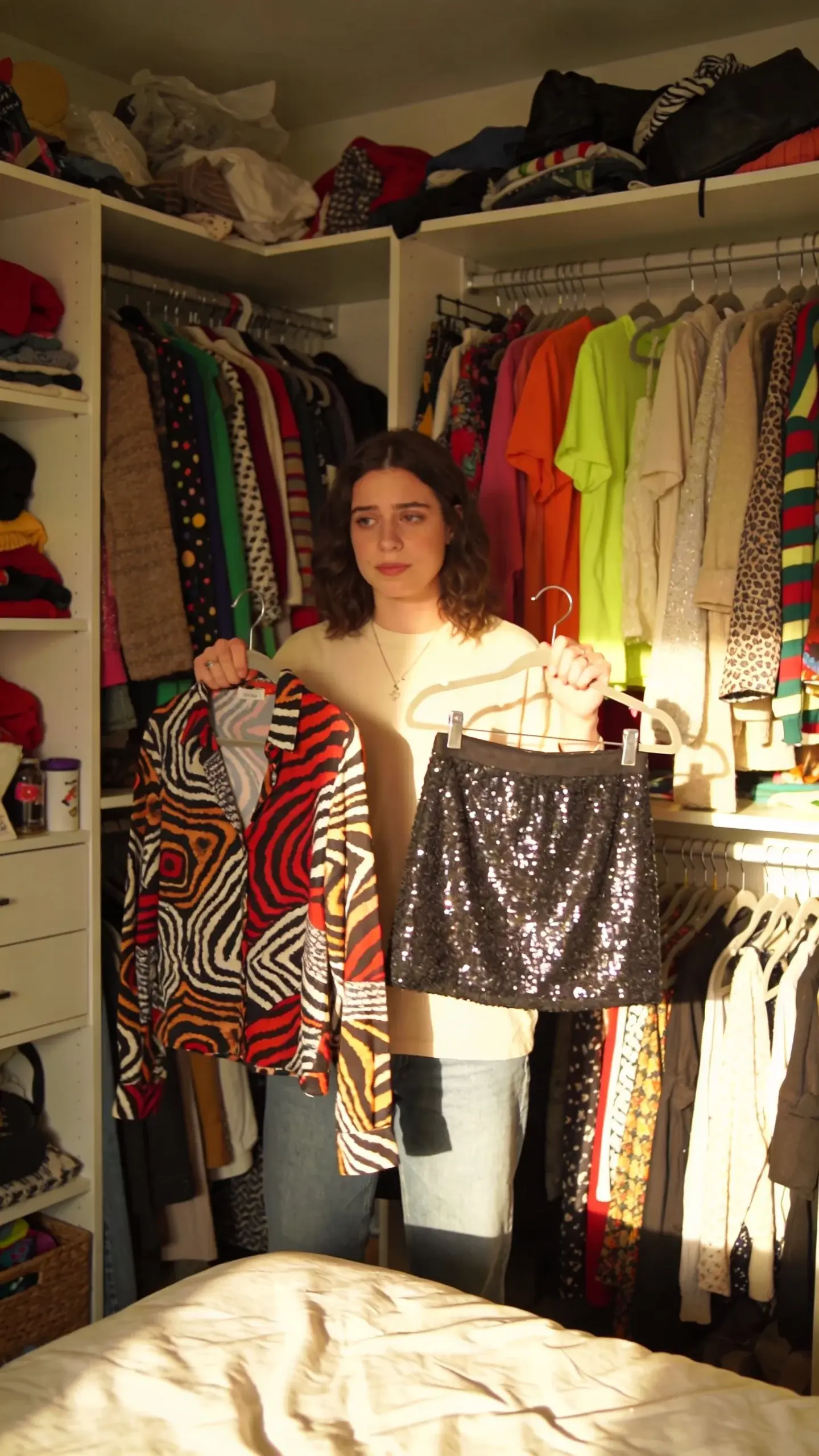 A cluttered, overstuffed closet with mismatched colorful garments on hangers and shelves, none in a cohesive palette; scattered bold prints, neon tops, sequined skirts, and random textures, with a person standing uncertainly in front holding two conflicting pieces, soft natural morning light, modern apartment bedroom.