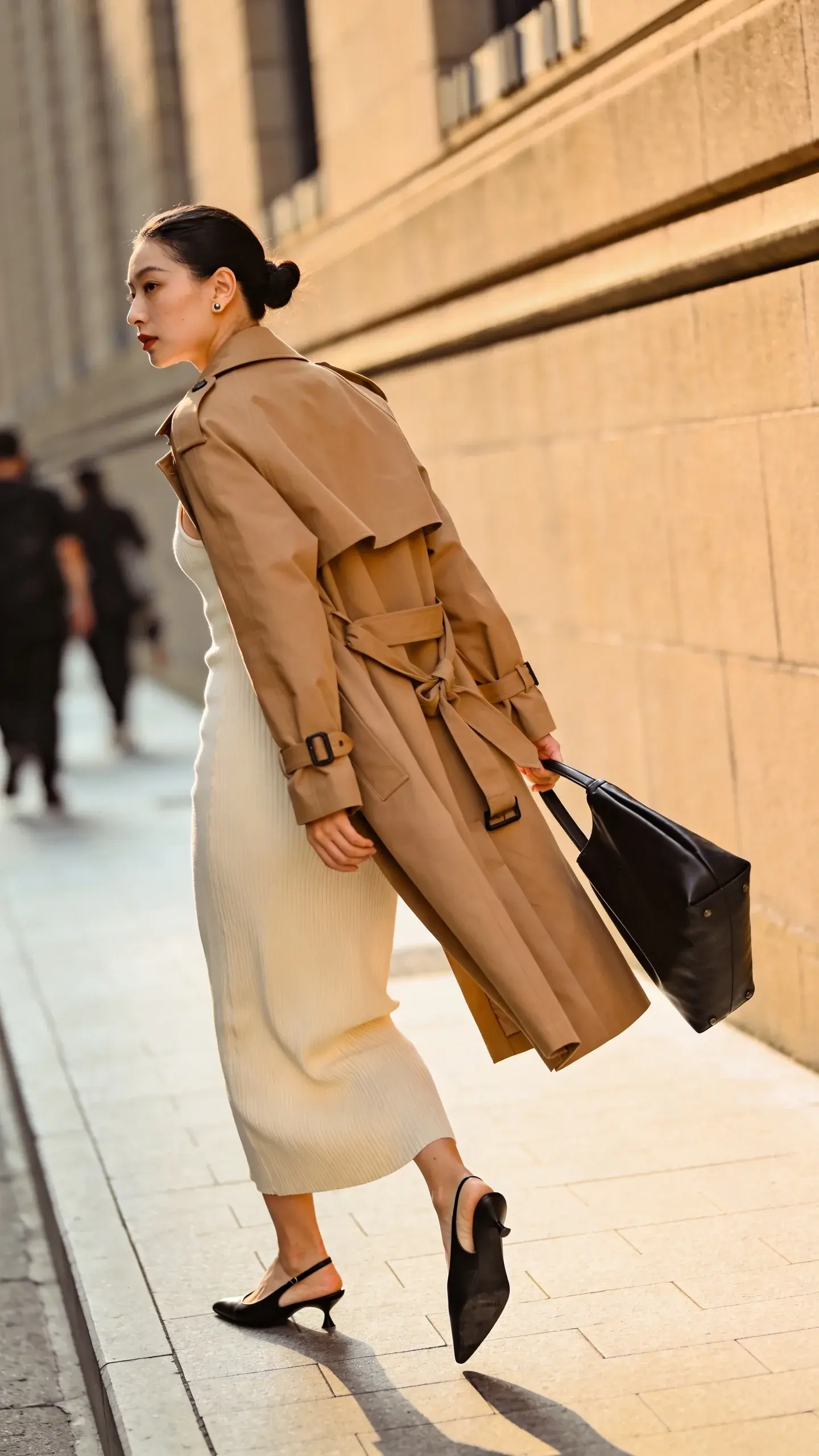 A minimalist, quiet-luxury street-style photograph of a woman wearing a camel trench coat with a classic belt knotted loosely at the back, layered open over a ribbed cream midi tank dress that has a bit of weight and skims without clinging; she pairs it with pointed black slingback flats, a structured black leather tote carried in-hand, and tiny minimal stud earrings. Clean, neutral palette and refined textures. Shot at golden hour on a softly lit city sidewalk with pale stone buildings and blurred pedestrians in the background; shallow depth of field, natural light creating gentle highlights on the trench’s drape and the ribbing of the dress. Composition frames her mid-stride, coat moving slightly, tote visible, shoes in focus. Realistic skin tones, subtle makeup, sleek low bun. 50mm lens look, editorial street-style photography, high resolution, no text.