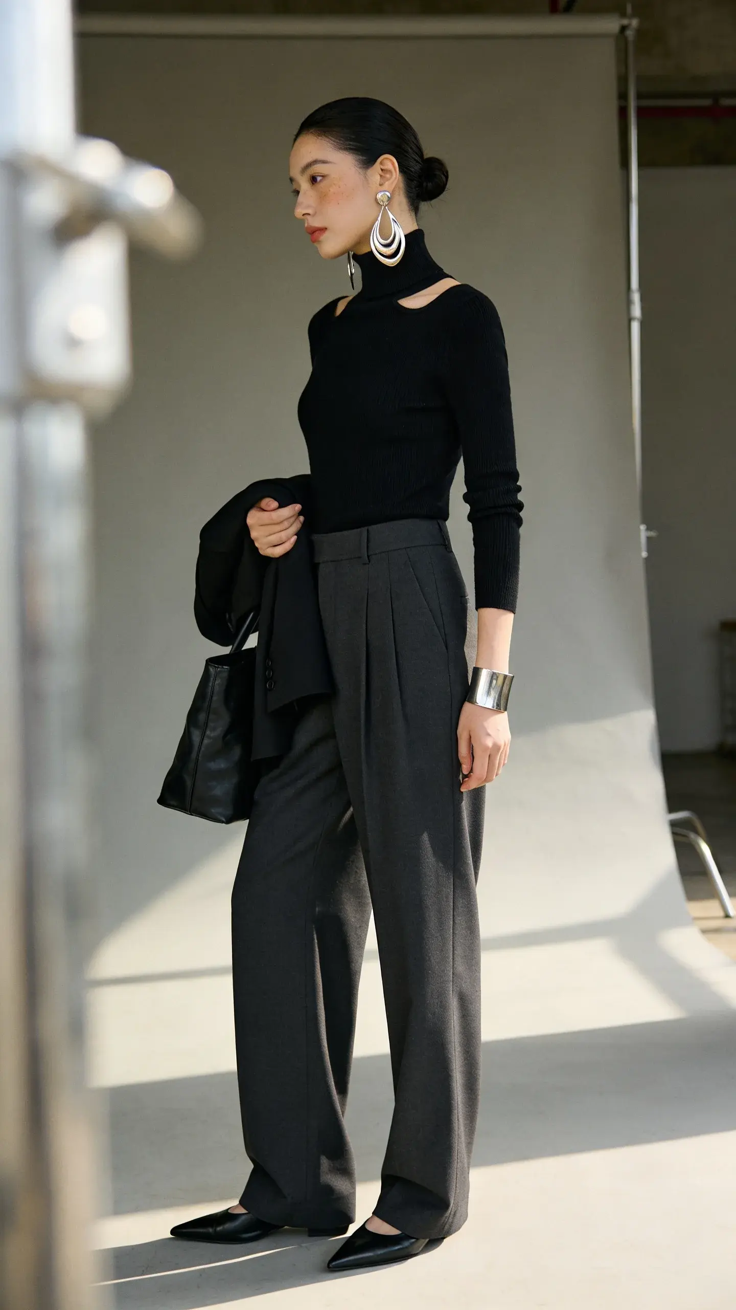 Elegant woman wearing sculptural earrings: a realistic, high-quality fashion photograph of a poised, modern woman in a minimal black turtleneck with a clean boatneck, tailored high-waisted wide-leg trousers in charcoal, and sleek black leather pointed-toe flats. The focal point is her bold yet refined sculptural metal earrings—polished silver abstract drop shapes with organic curves and clean lines—paired with a slim matching silver cuff bracelet. Hair is pulled into a smooth low bun to balance scale and showcase the earrings; makeup is fresh with subtle highlight and soft nude lip. She stands three-quarter profile against a softly lit neutral studio backdrop (warm gray), natural daylight look with gentle shadows to emphasize texture and gleam (not glitter). Composition frames from mid-thigh up, drawing the eye up to the earrings; minimal styling—no necklace—to keep the neckline clean. Add a structured black leather tote resting at her side and a hint of a tailored black blazer draped over her forearm to suggest versatility. Photography style: editorial street-meets-studio, shallow depth of field, crisp detail on metal surfaces and knit texture, true-to-life color, high dynamic range, 50mm lens perspective.