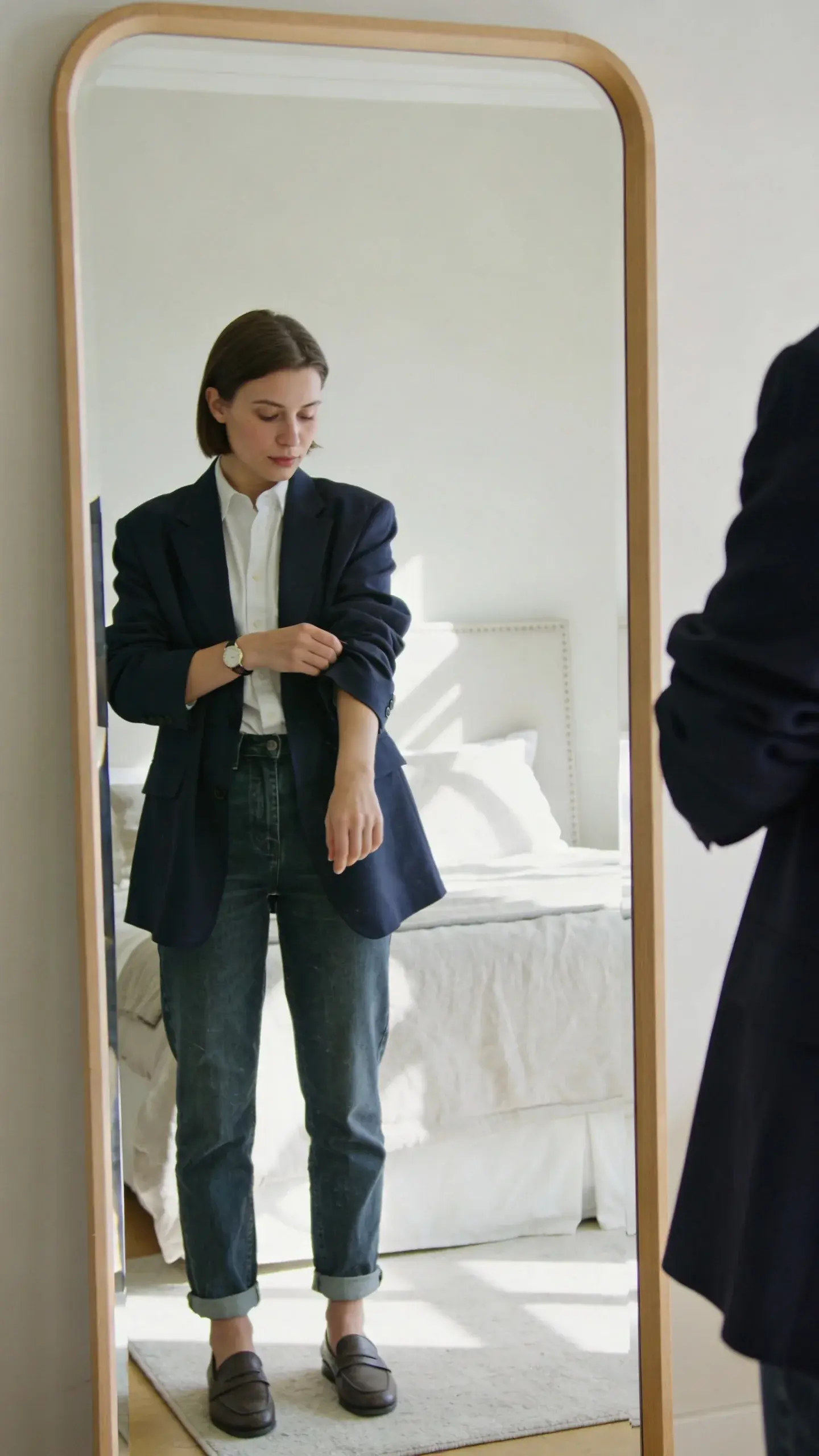 A well-lit bedroom scene with a woman standing in front of a mirror, folding the sleeves of a slightly oversized navy blazer to mid-forearm to reveal a slim watch and a sliver of wrist; she’s wearing a white button-down half-tucked into straight-leg medium-wash jeans, cuffs rolled to hit just at the ankle bone, paired with simple leather loafers; neutral palette, soft morning light, no text.