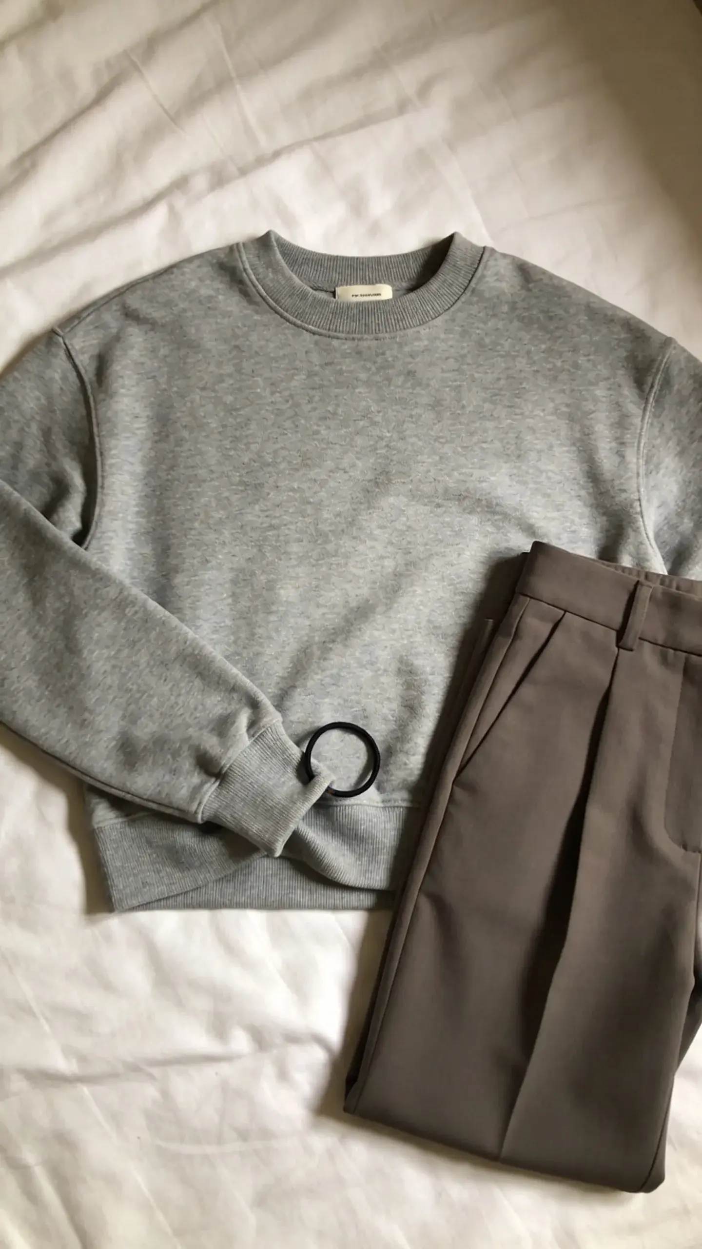A step-by-step style still-life: a grey crewneck sweater laid flat on a clean bed, a small black hair tie looped around a pinch of the sweater hem and tucked under to create a cropped effect; include a pair of high-waisted trousers beside it; overhead shot, soft shadows, no text.