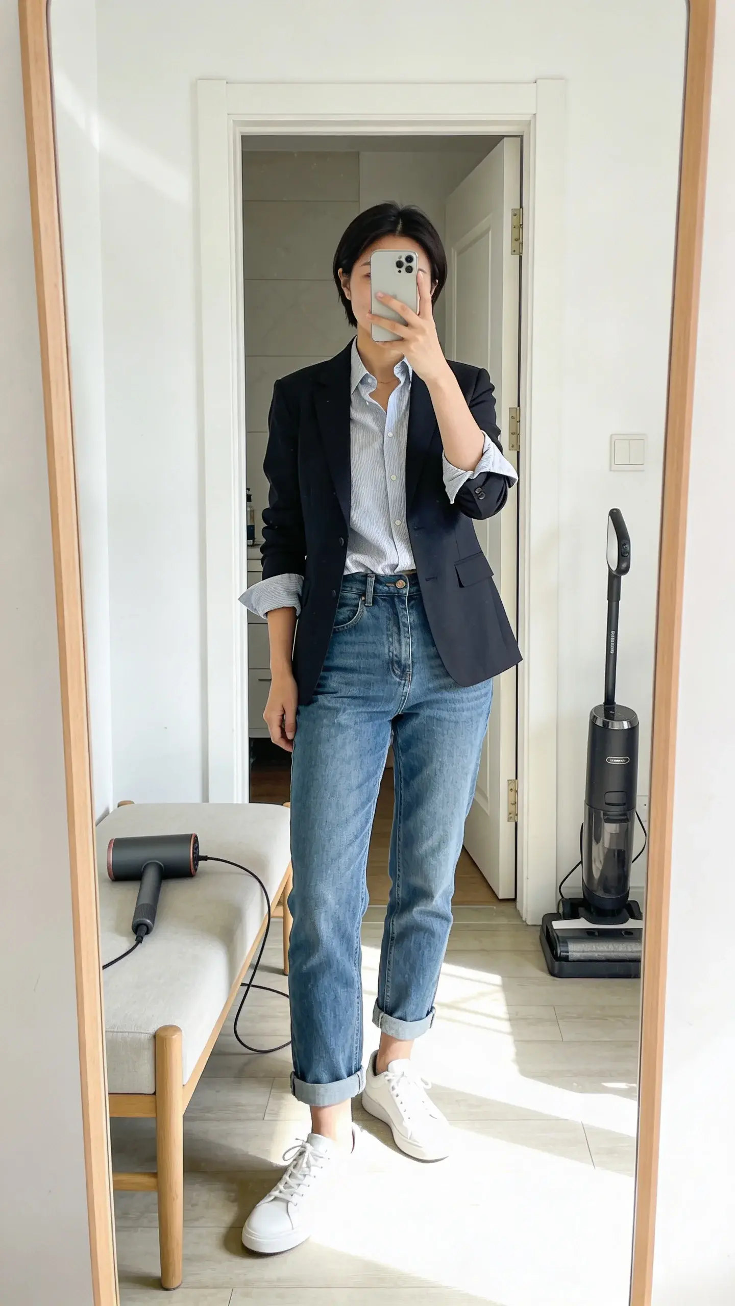 A full-length mirror selfie style composition (no text on mirror) showing cuffed straight-leg jeans hitting the ankle bone, paired with clean white sneakers; the shirt is half-tucked, blazer sleeves rolled; a lint roller and steamer rest on a bench in the background; bright, airy entryway, no text.