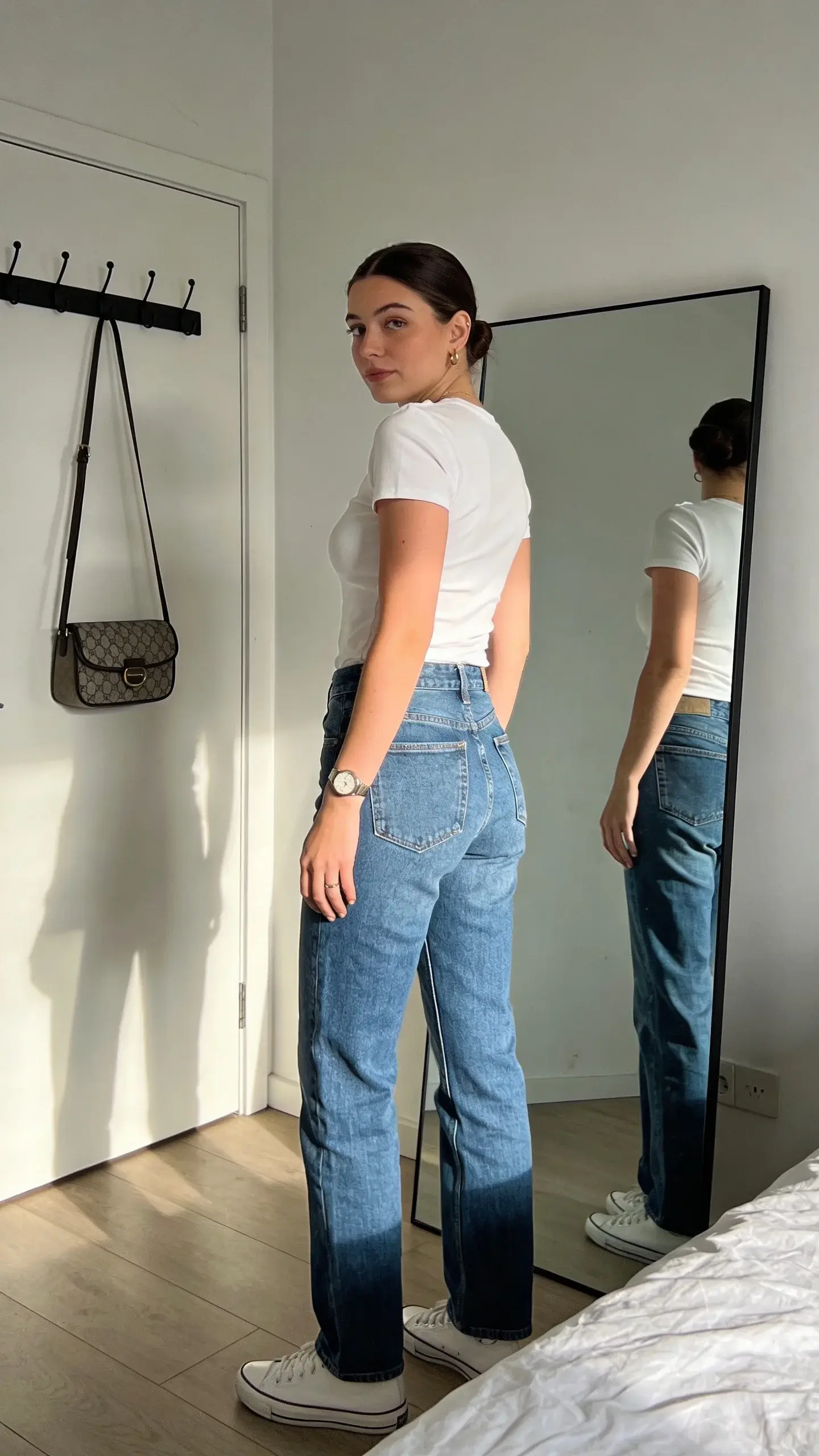 A modern, sunlit bedroom with a full-length mirror: a woman in her late 20s wearing a plain white fitted tee and mid-rise straight-leg blue jeans stands relaxed, turning slightly to the side to show shoulder seams aligned perfectly with her shoulder bones; minimal jewelry (small hoops, simple watch), sleek low bun, clean white sneakers, and a structured crossbody bag on a hook in the background; neutral palette, soft natural light, no text.